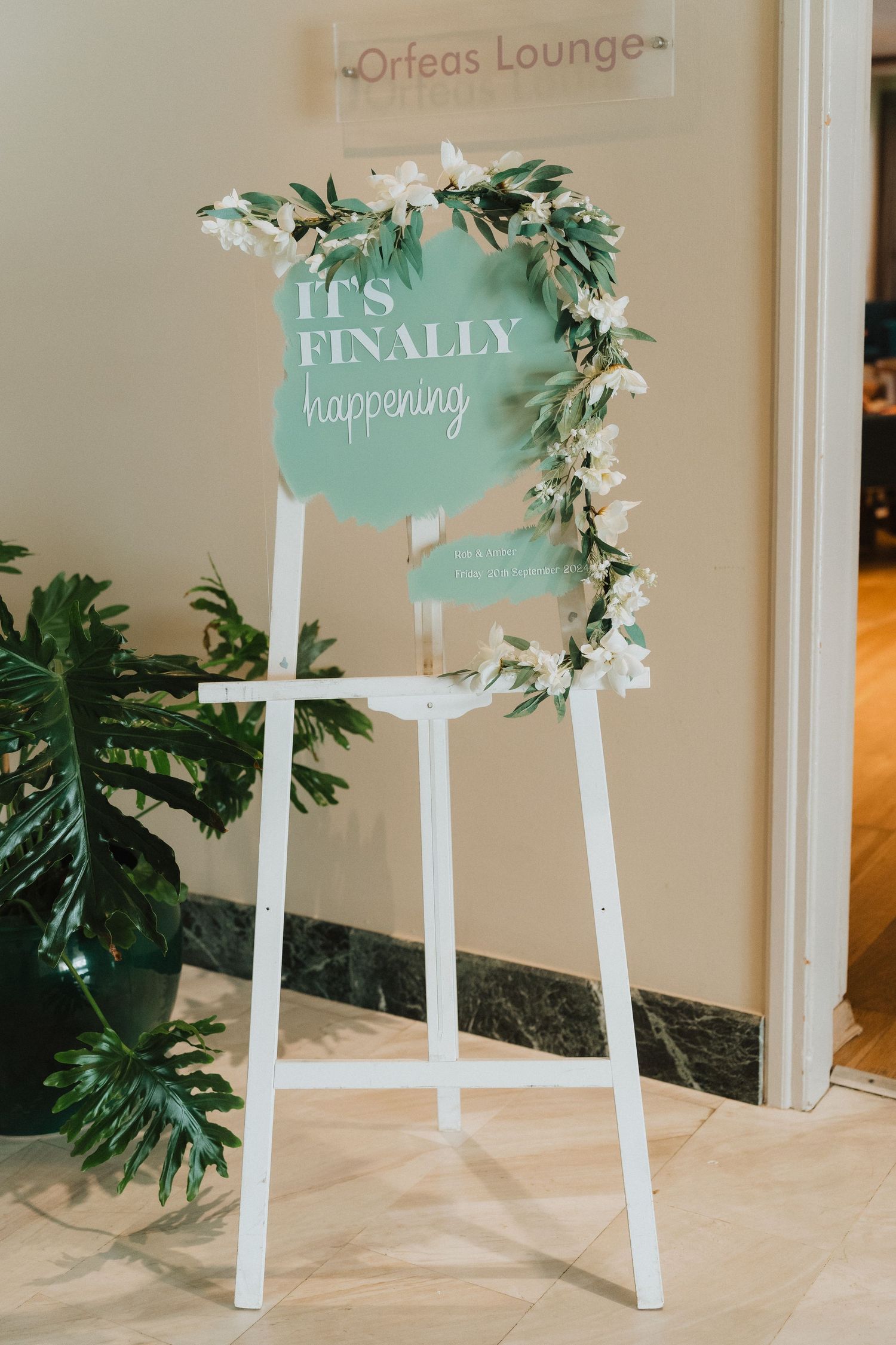A mint green wedding sign with 'It's Finally Happening' text decorated with white floral garland on a white easel stand.