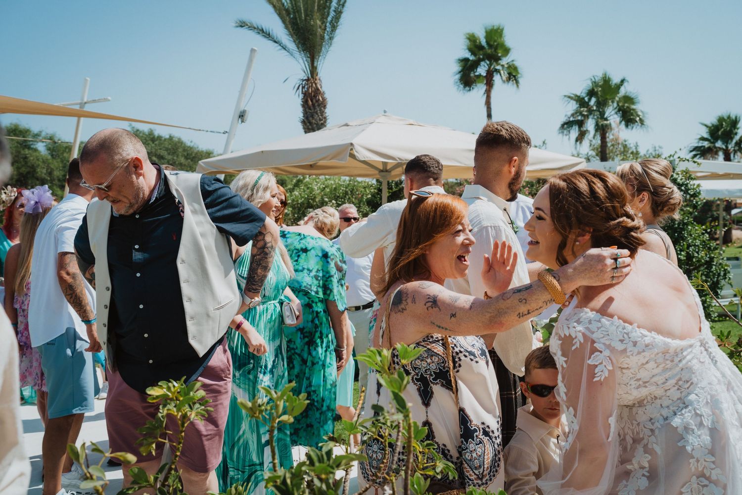 Wedding guests mingling outdoors at a sunny tropical venue with palm trees and shade canopies.
