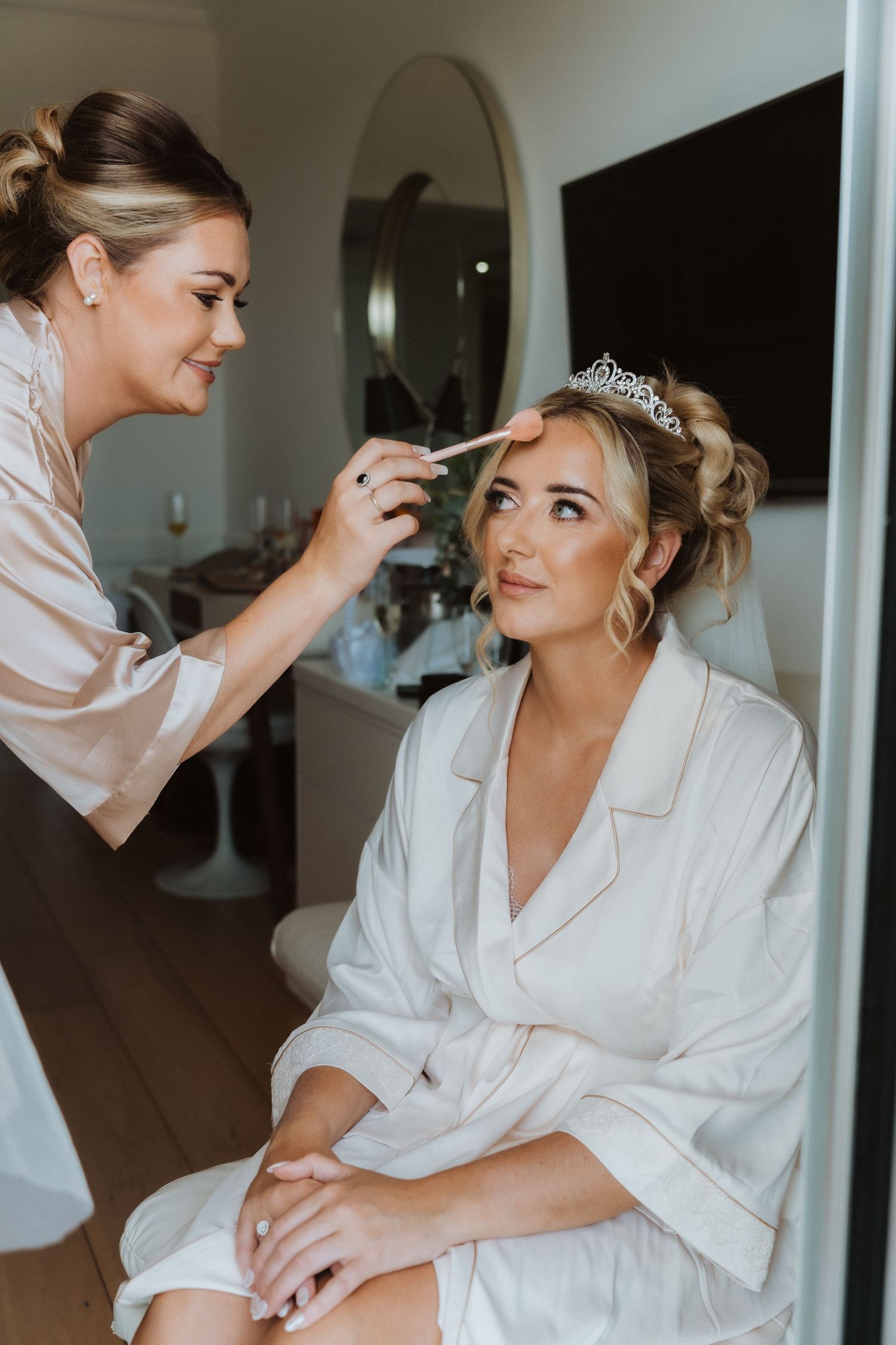 Makeup artist applies final touches to a bride wearing a white robe and tiara on her wedding morning.