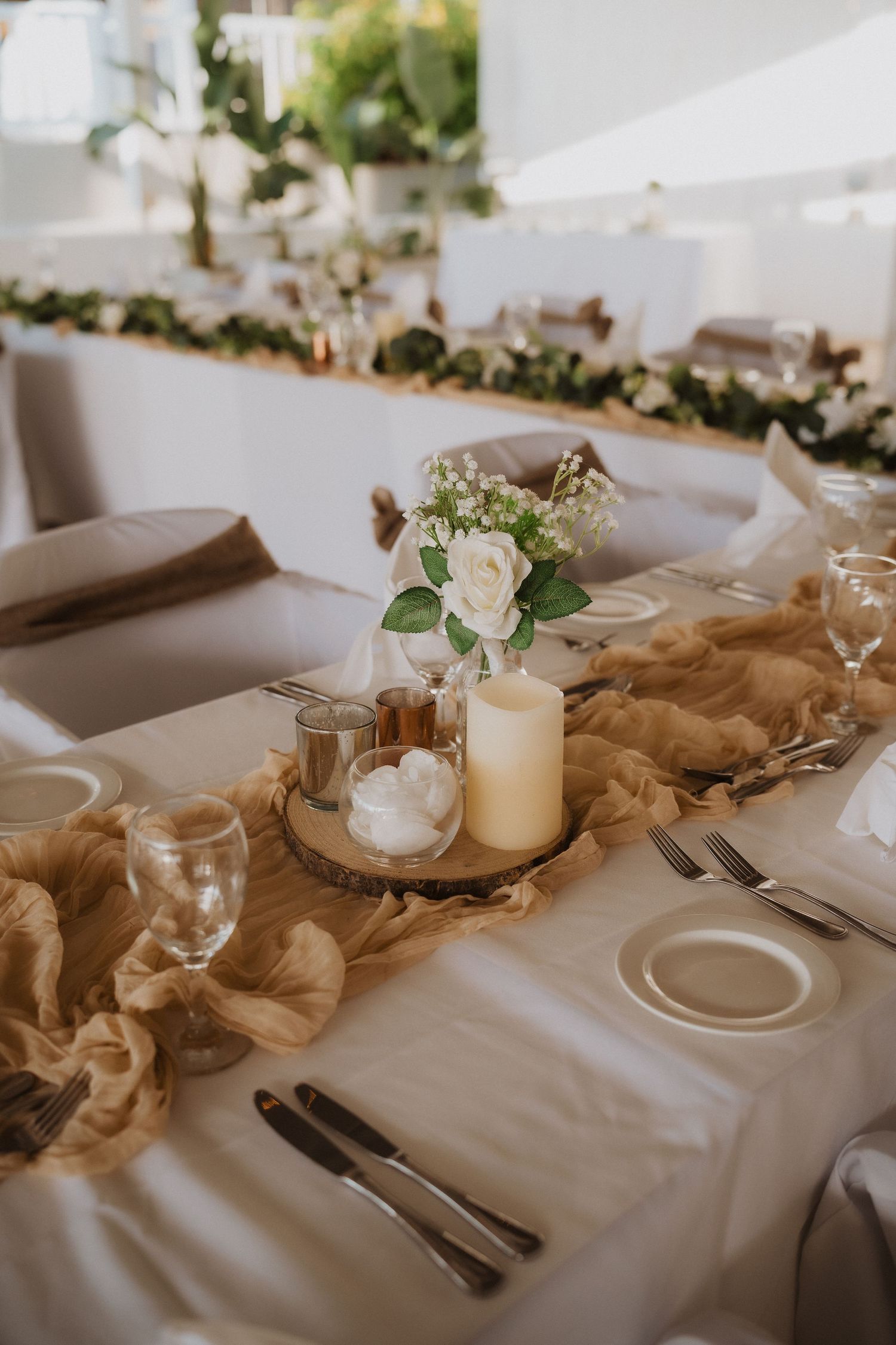 Rustic wedding table setting with candles, white roses, and flowing tan fabric runner.