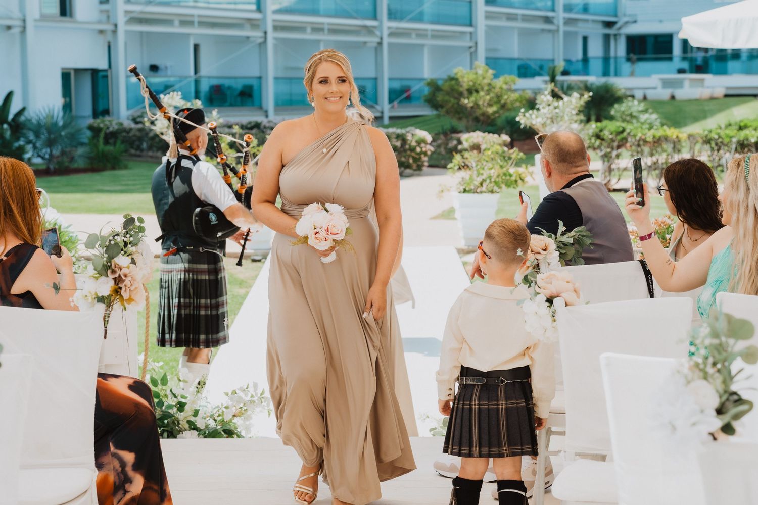 Outdoor wedding ceremony with bagpiper and attendants walking down decorated aisle.