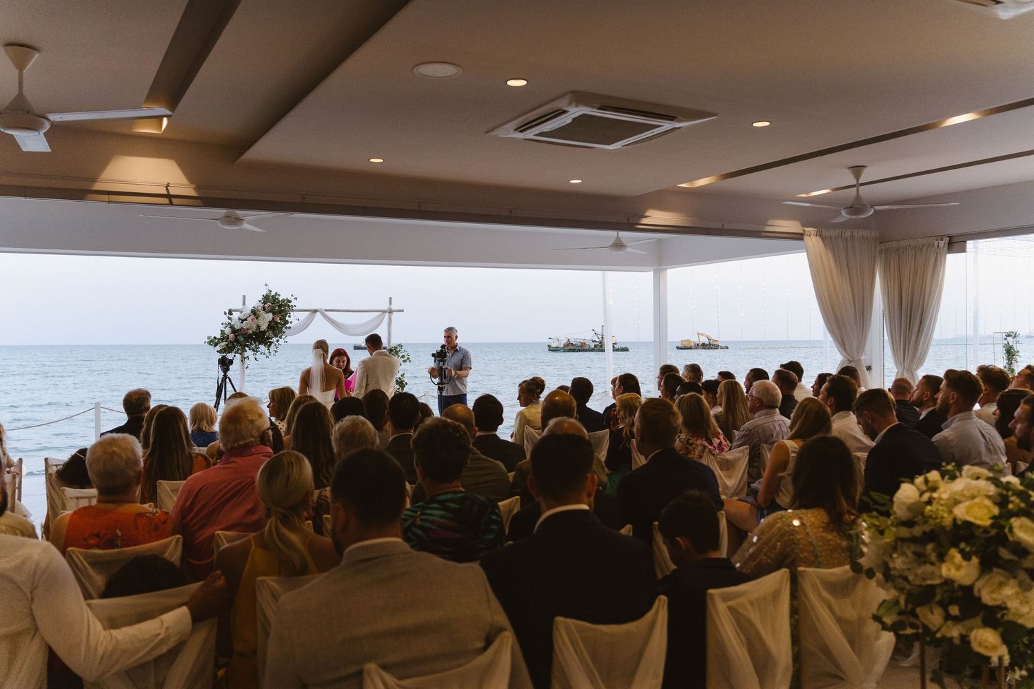 Scenic beach wedding ceremony with guests seated under covered pavilion overlooking ocean.