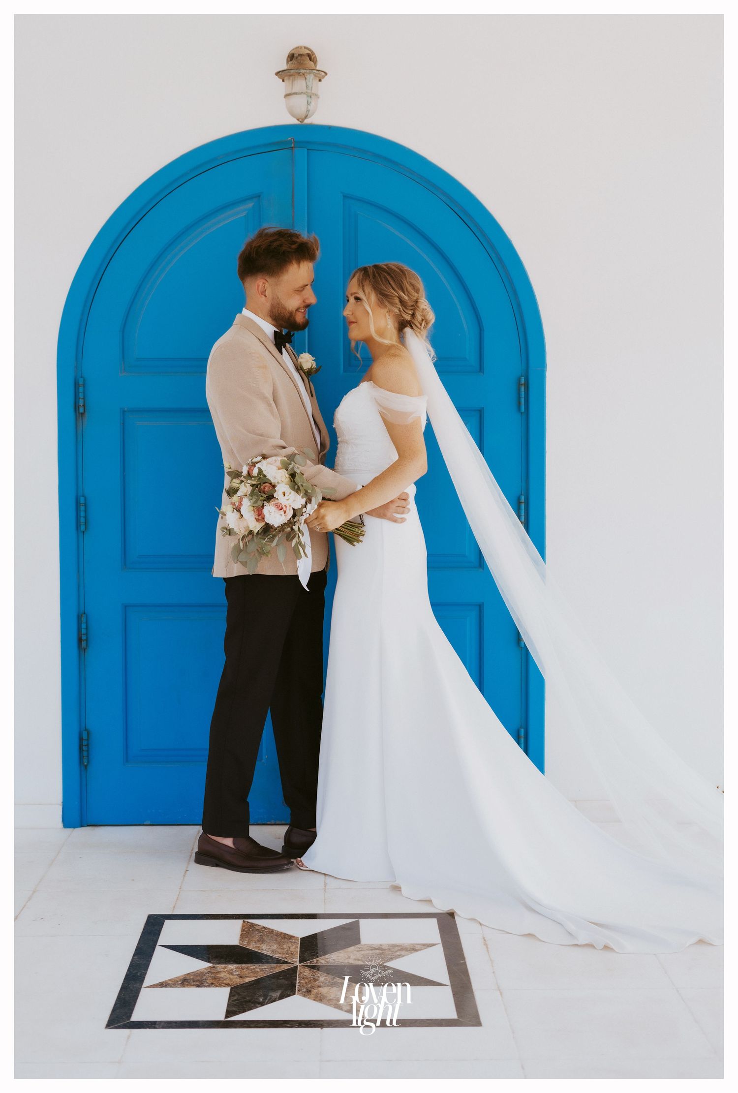 Wedding couple poses in front of striking blue arched door holding floral bouquet.