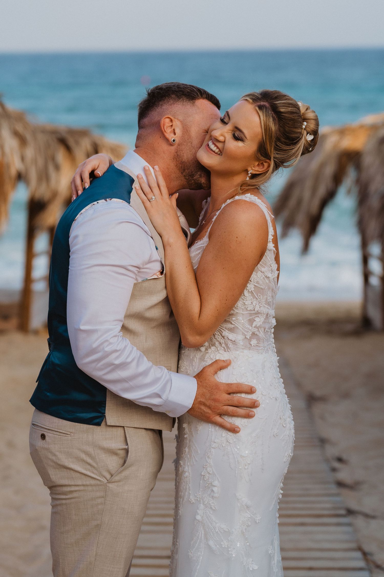 A romantic couple shares intimate kisses on a tropical beach with thatched umbrellas and turquoise waters in the background.