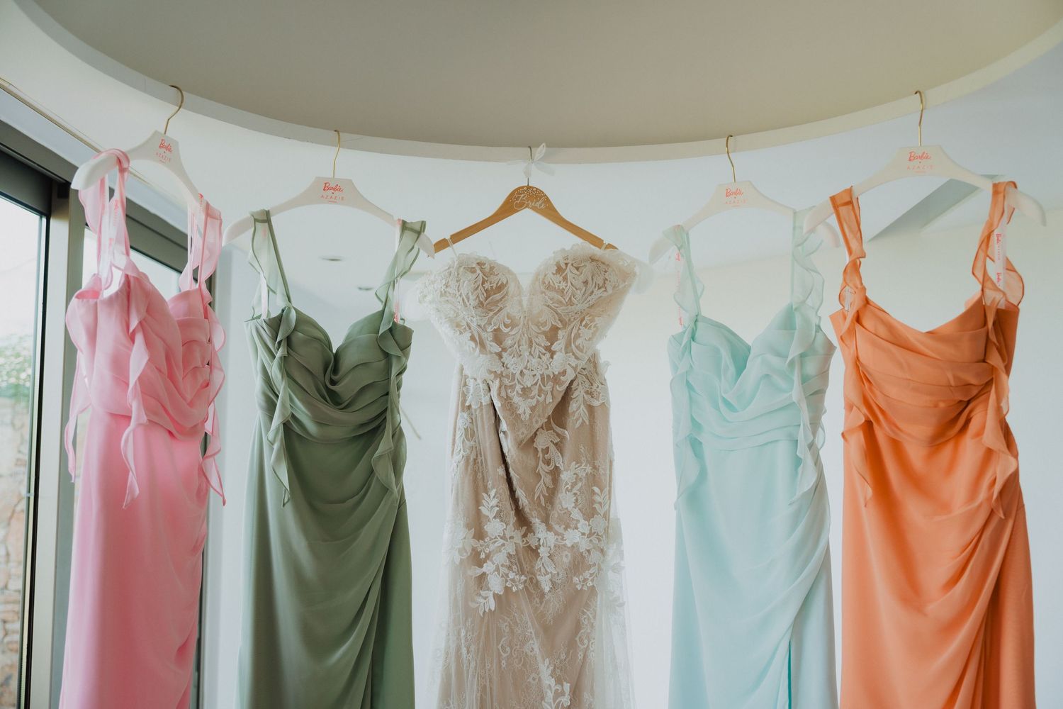 A row of pastel-colored bridesmaid dresses displayed on hangers against a white wall.