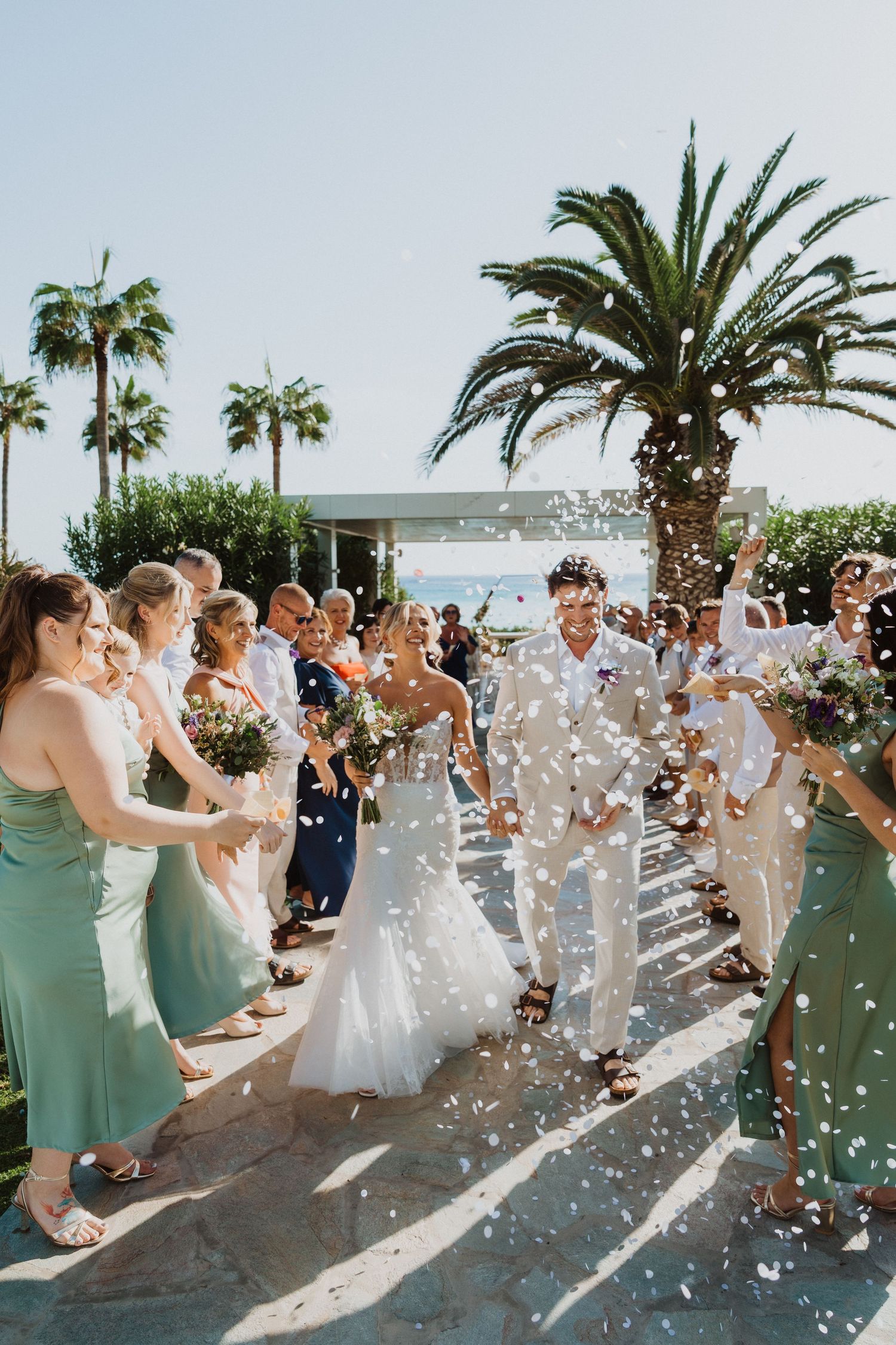 Sunlit wedding celebration with confetti being tossed by guests near tropical palm trees.