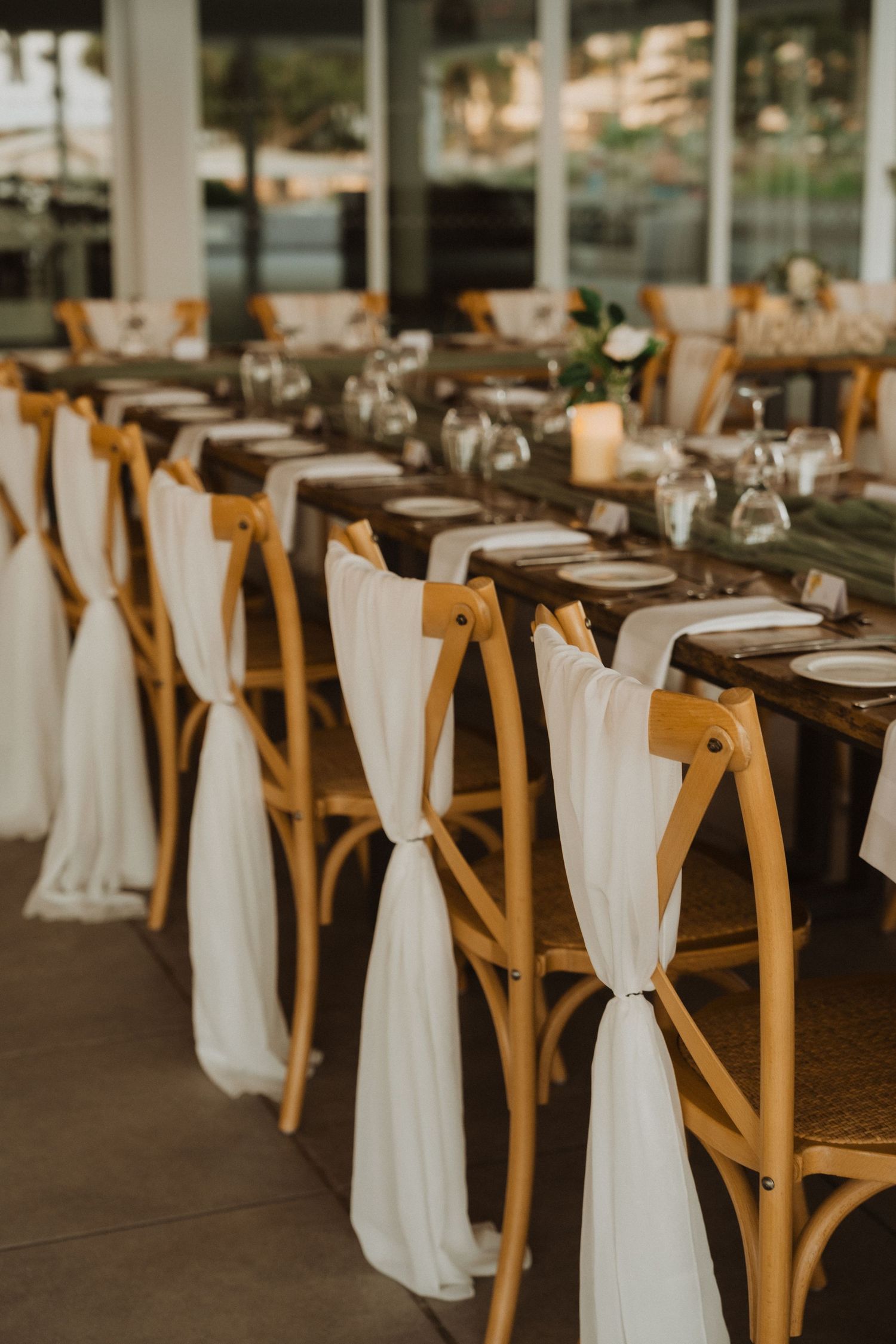 Close-up of wooden crossback chairs adorned with white fabric sashes at a wedding reception.