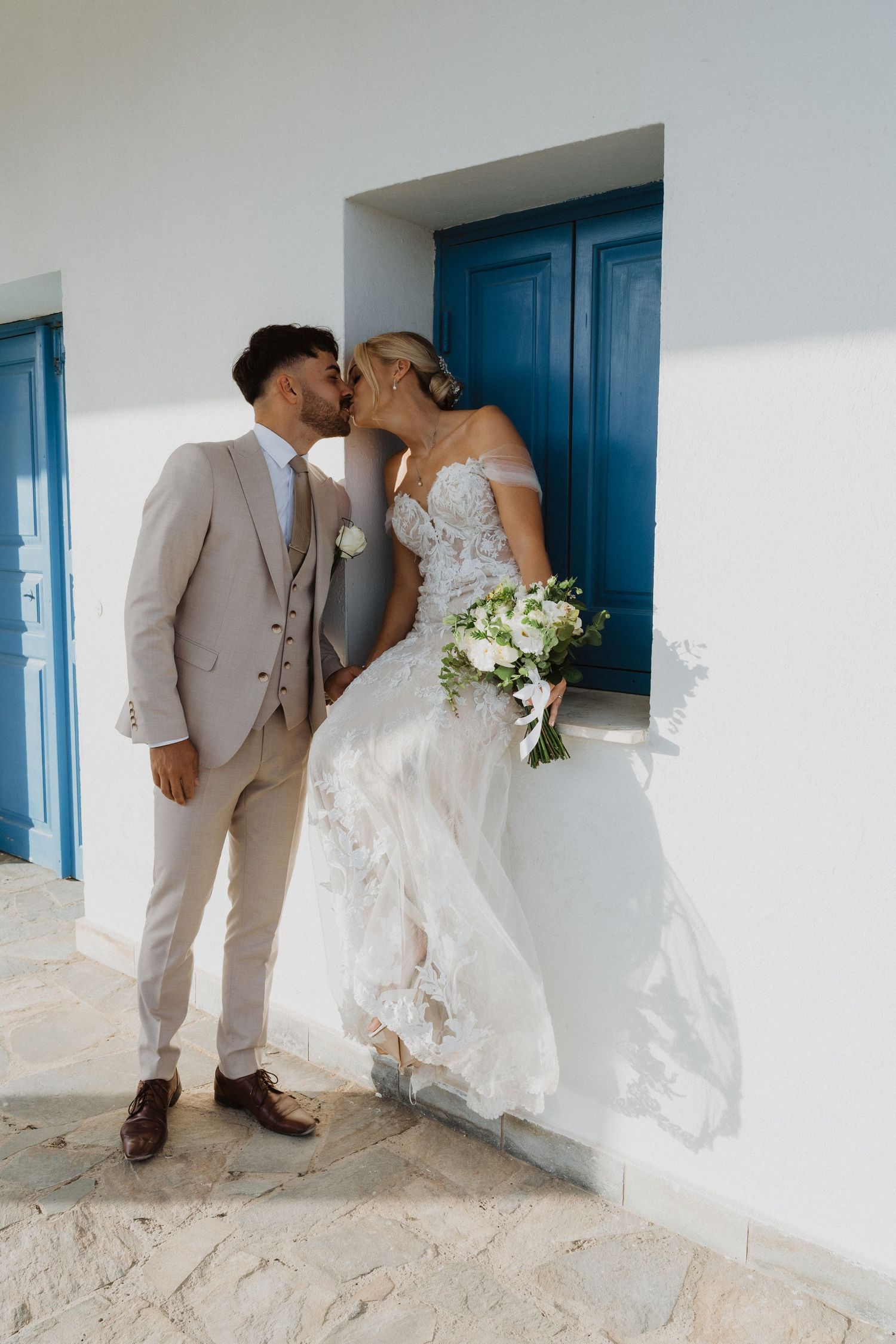 Newlyweds share intimate moments along a white Mediterranean building with blue accents.