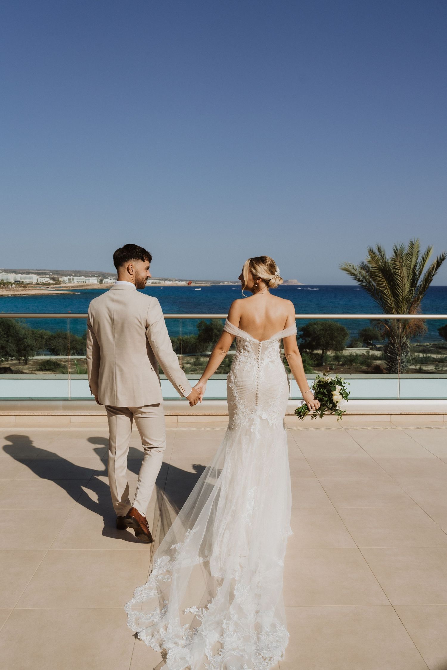 Wedding couple holding hands on a beachfront terrace overlooking the Mediterranean Sea.