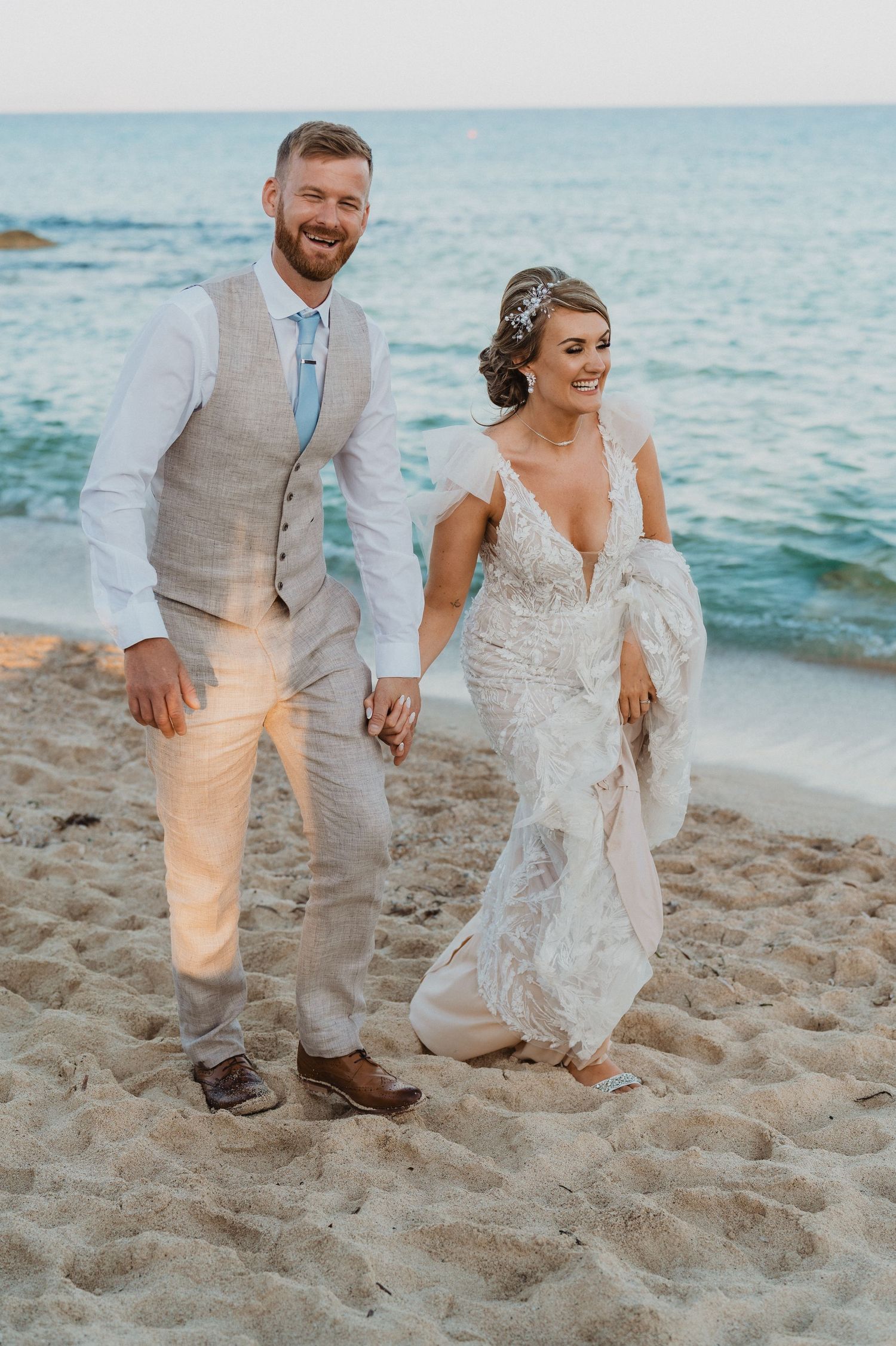 Newlyweds stroll hand in hand on tropical beach with turquoise water backdrop.