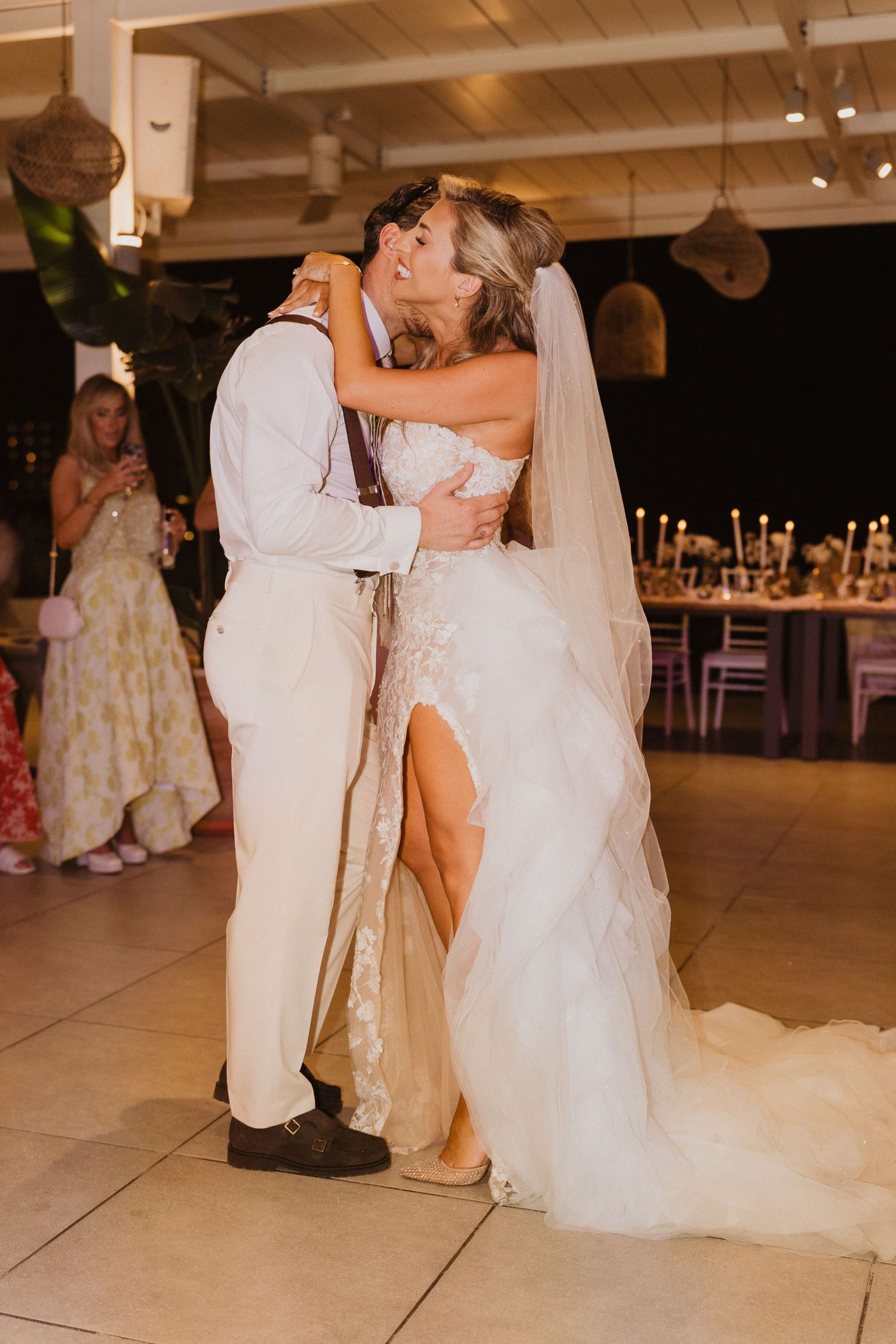A couple shares their first dance at their wedding reception in an elegant venue with candlelit tables in the background.