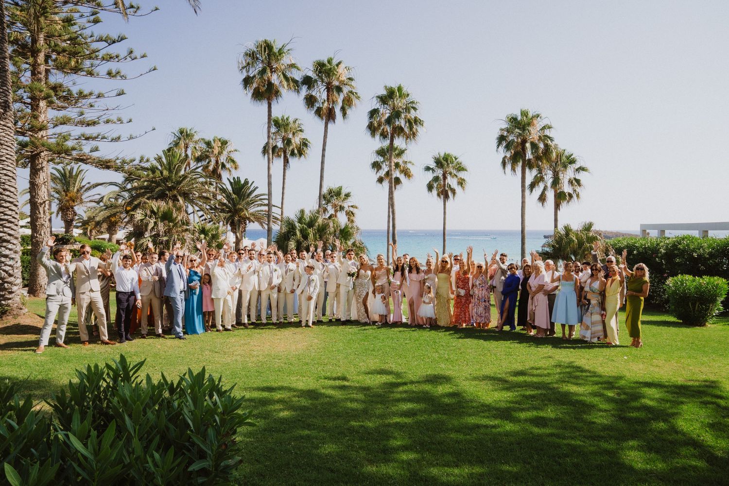 Large wedding party gathered on a lush green lawn with tall palm trees and ocean views in the background.