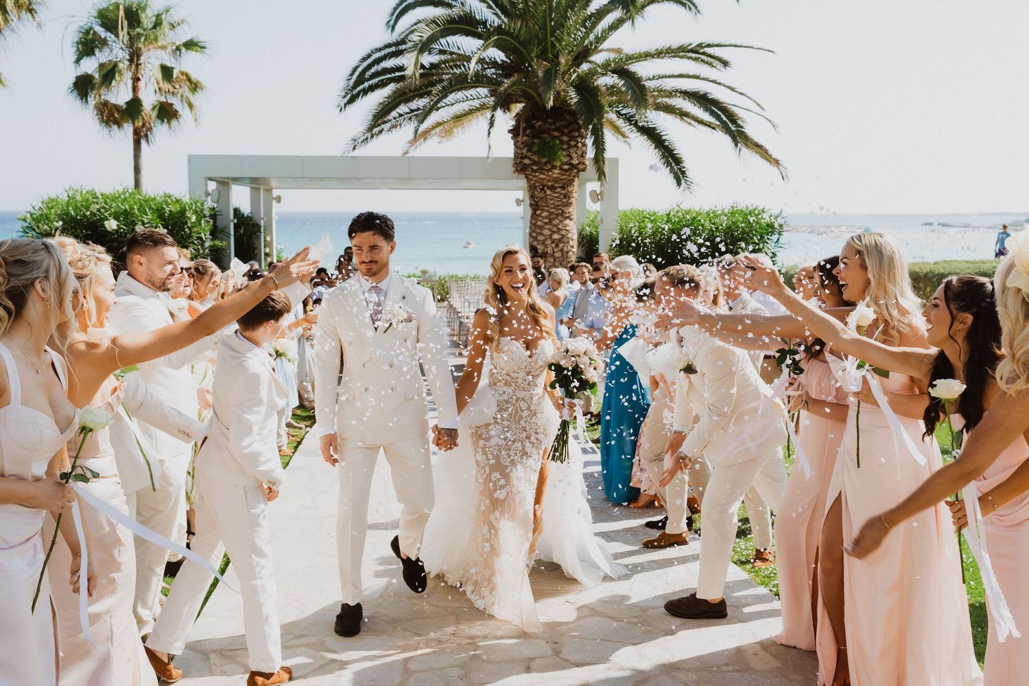 Wedding party celebrates on a sunny terrace with palm trees and ocean views as guests form an archway for newlyweds.