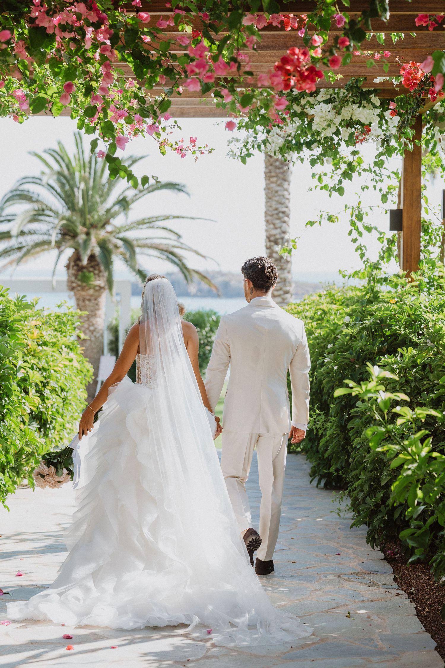 A couple walks along a scenic garden pathway lined with pink bougainvillea and palm trees on their wedding day.