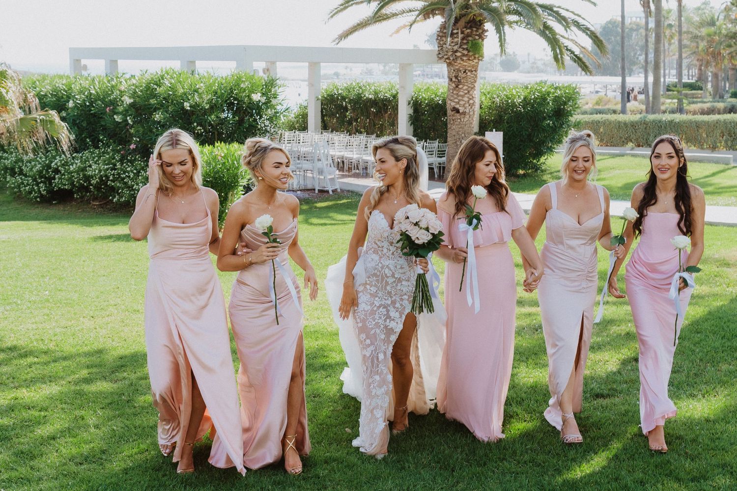 Bridesmaids in flowing pink dresses walk together and laugh on a grassy lawn at an outdoor wedding venue.