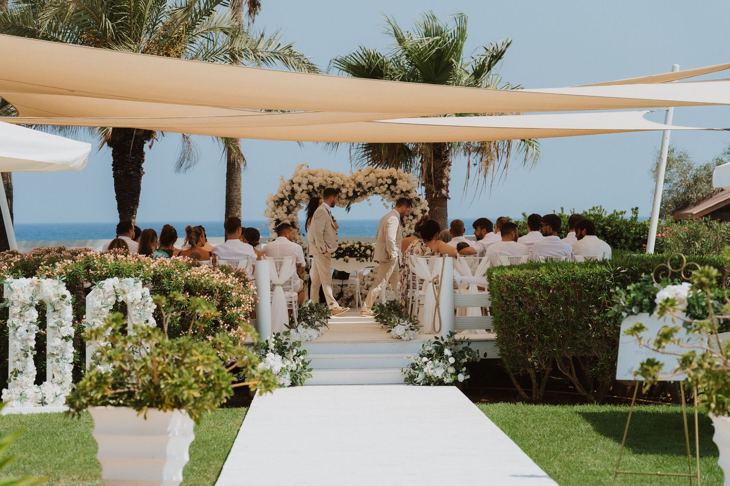 Elegant outdoor wedding ceremony on lush green lawn with white aisle, floral arch, and ocean view under cream shade sails.
