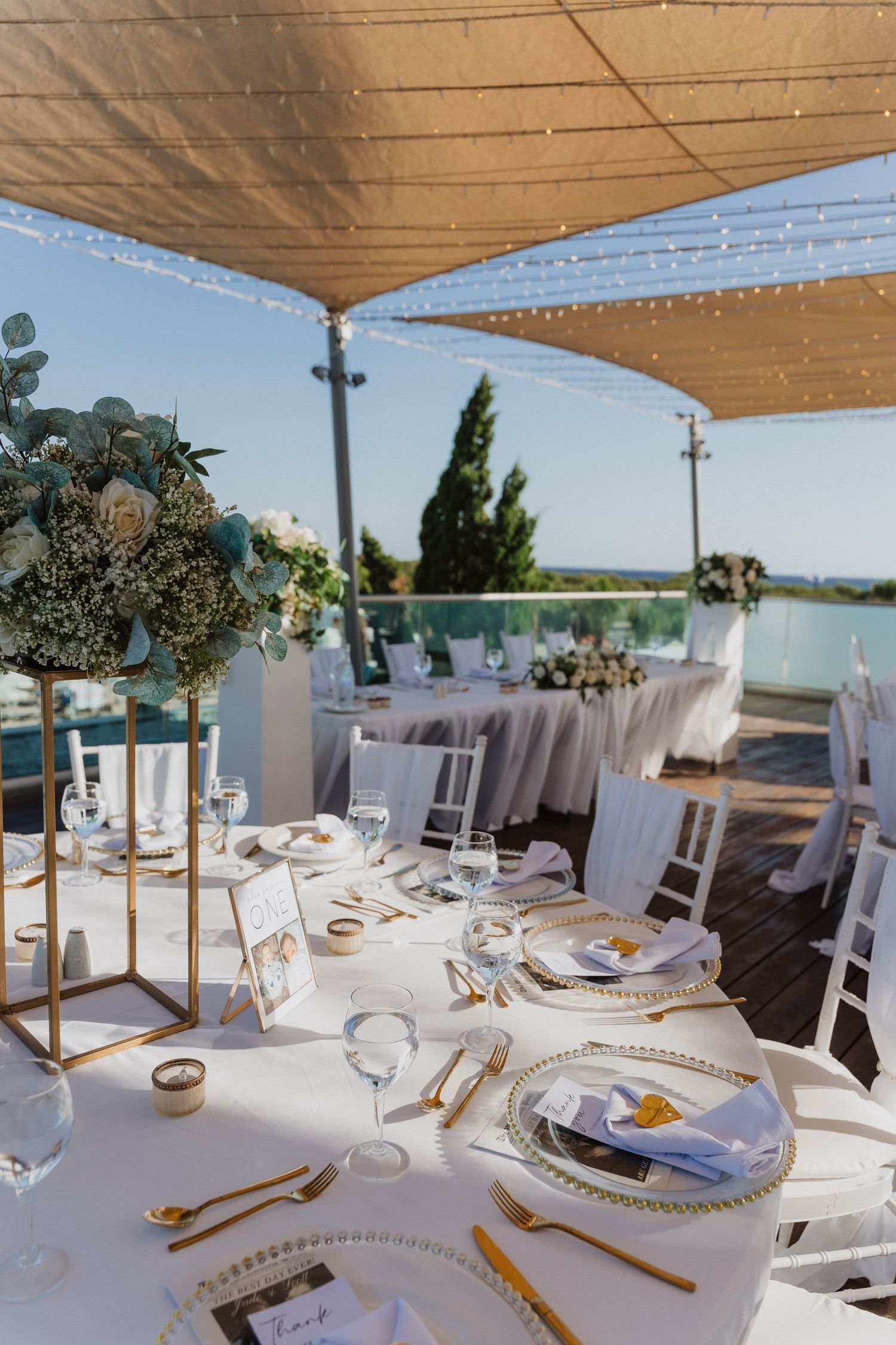 Elegant outdoor wedding reception setup with white tables, gold accents and floral centerpieces under beige canopy by the sea.