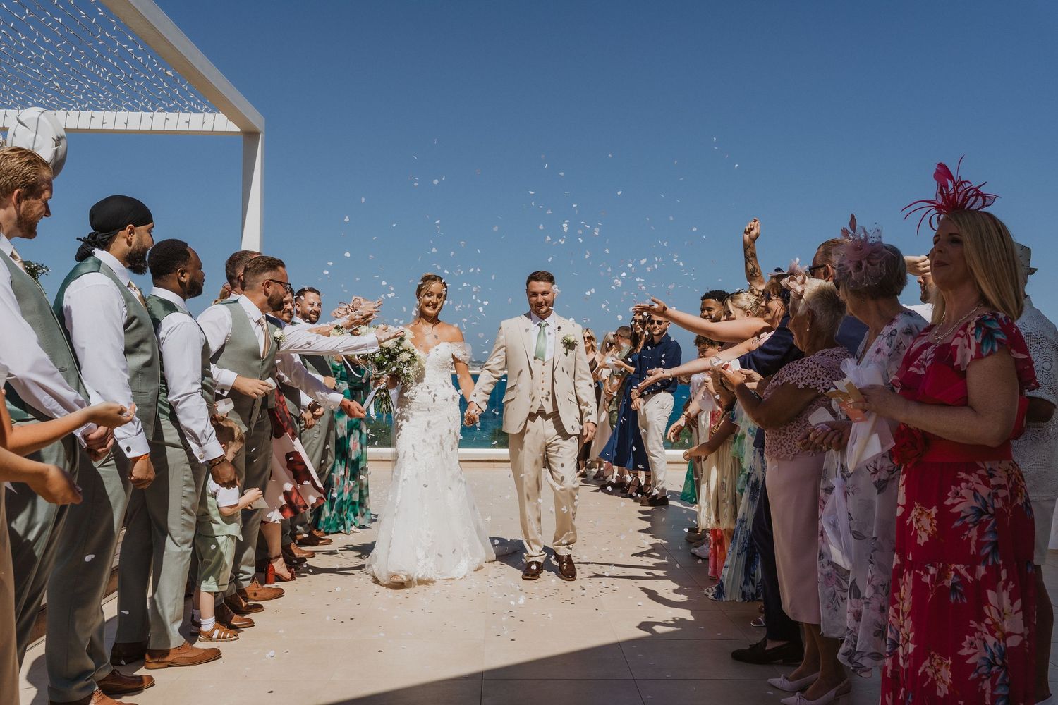 Wedding guests toss flower petals and celebrate along a beachside walkway during a sunny outdoor ceremony.