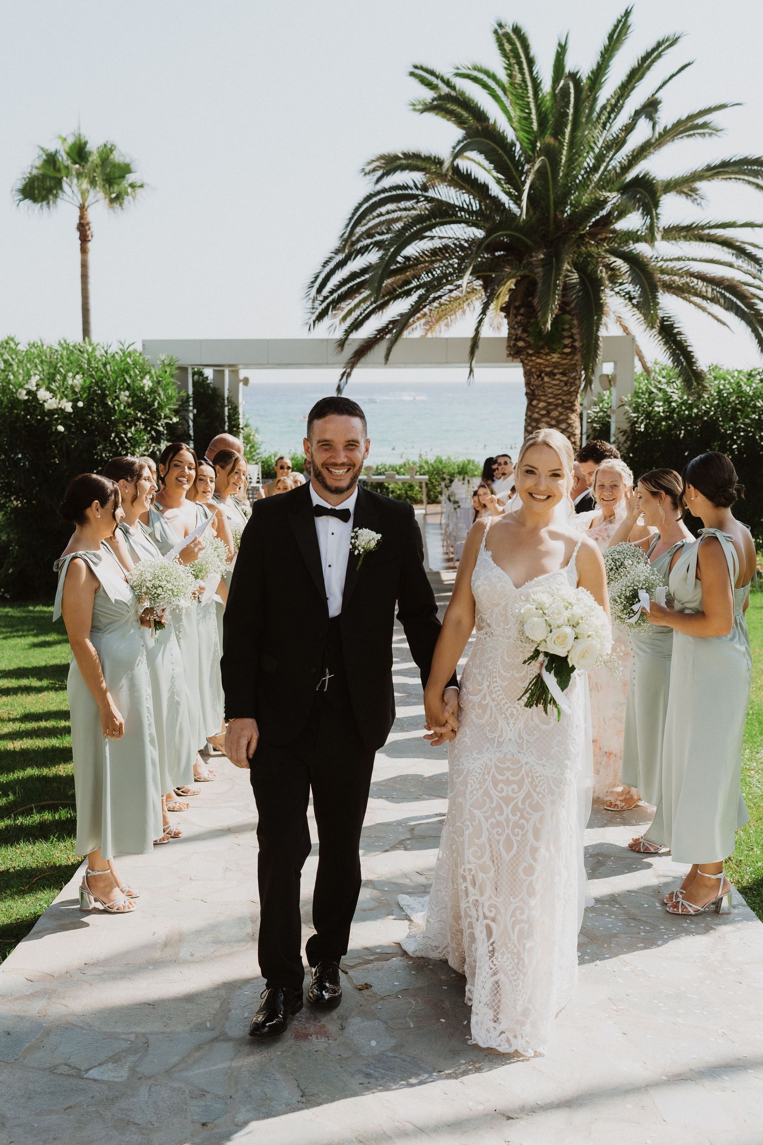 Wedding party walks along a palm-lined pathway at a sunny outdoor venue.