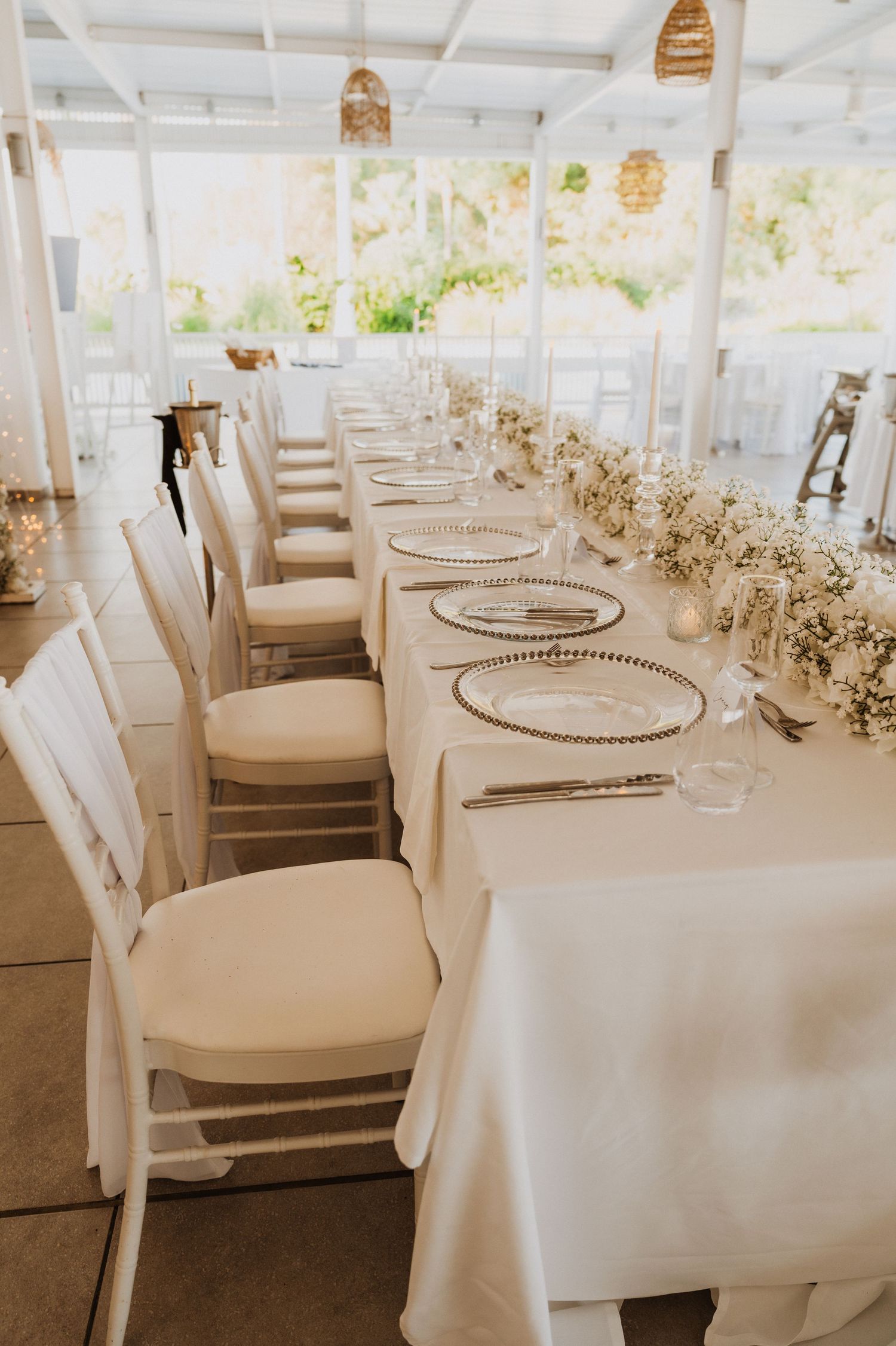 Elegant wedding reception table setting with white chairs and linens decorated with delicate floral arrangements.