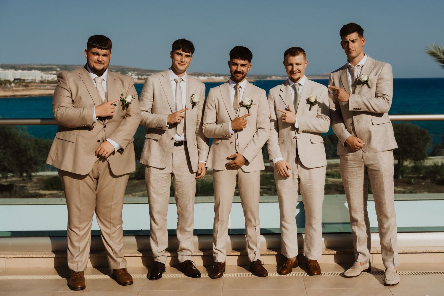 Groomsmen in beige suits pose on oceanfront terrace.