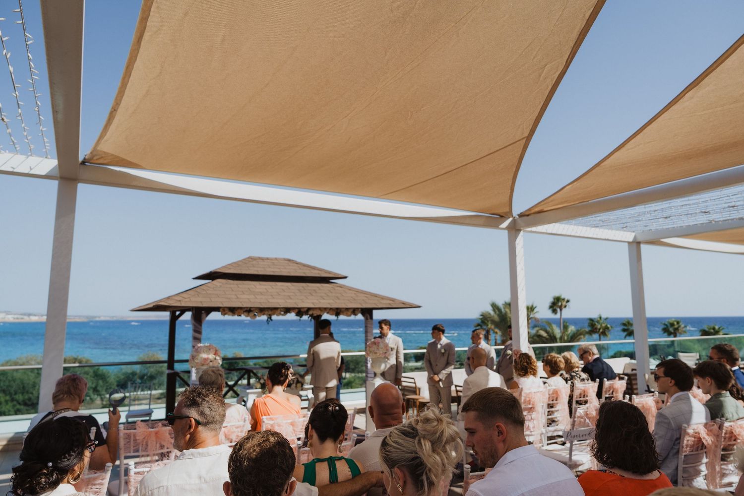 Guests seated under beige canopies at an outdoor beach wedding ceremony with ocean views.