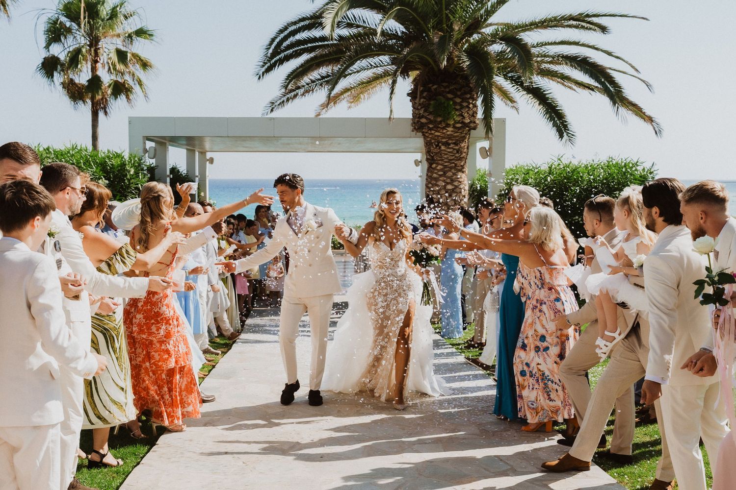 Wedding guests toss flower petals at a celebration under palm trees by the ocean on a sunny day.