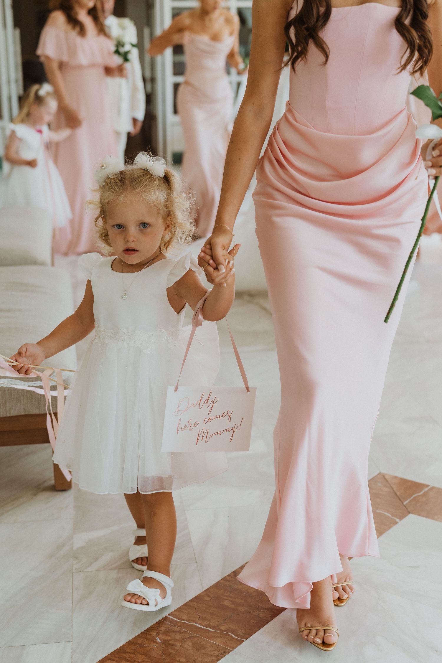 Wedding party members in soft pink dresses and white sandals stand together holding single-stem flowers.