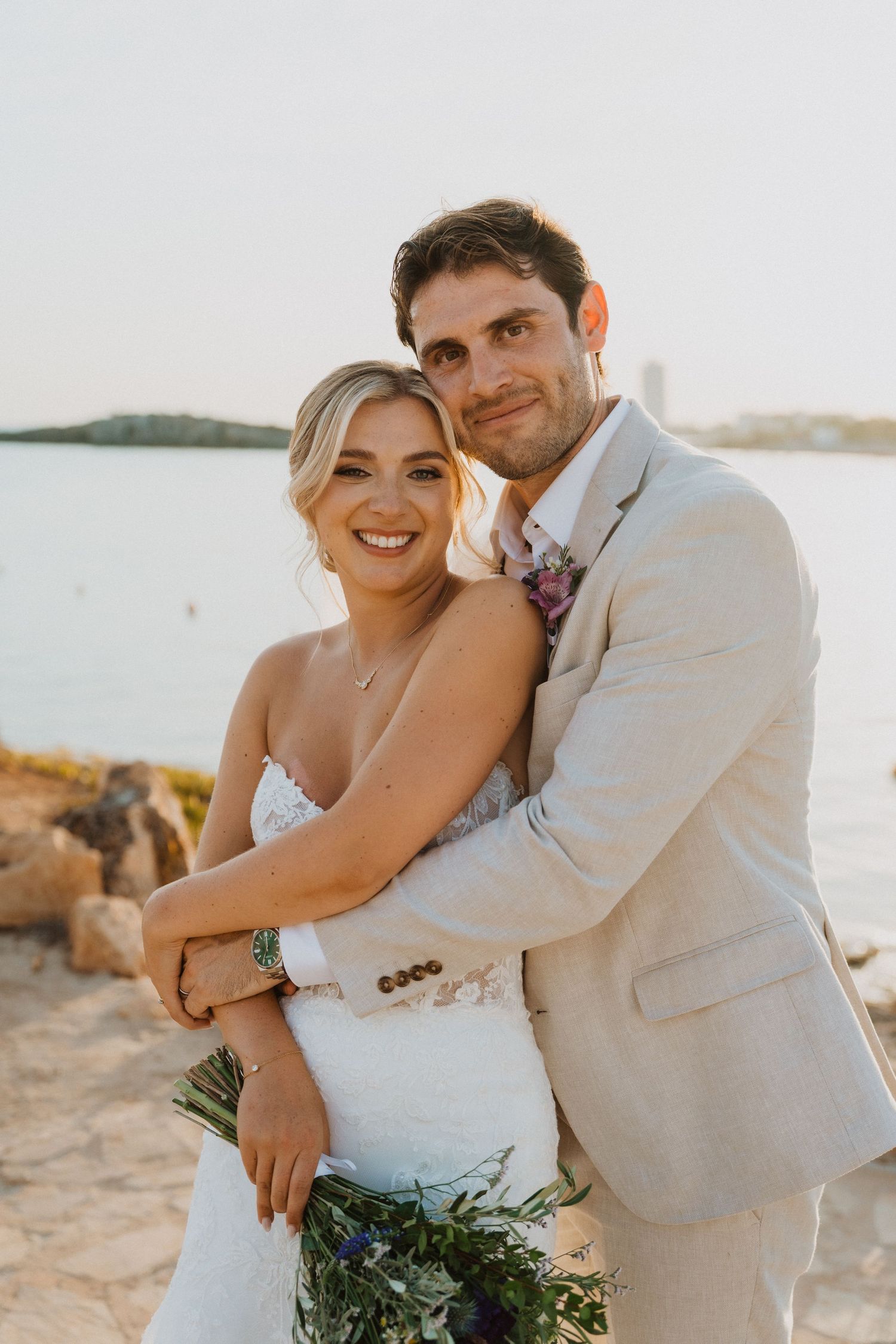 A romantic wedding portrait of a couple embracing on a coastal cliff at golden hour.