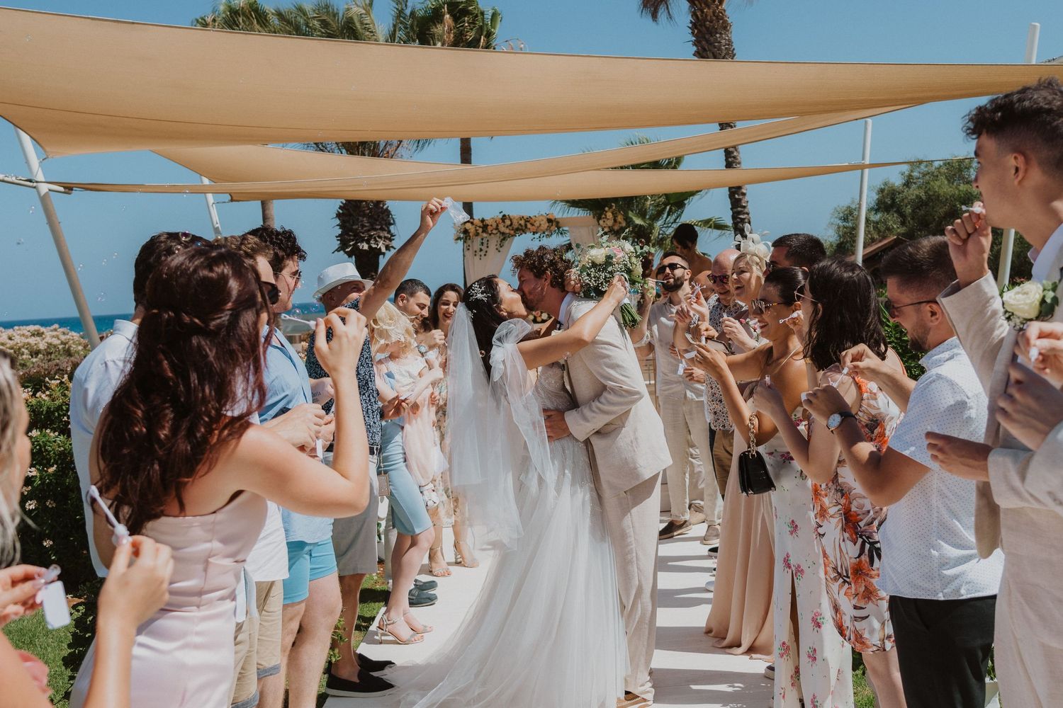 Wedding guests celebrate on a beachfront venue covered by tan shade sails throwing confetti into the air.