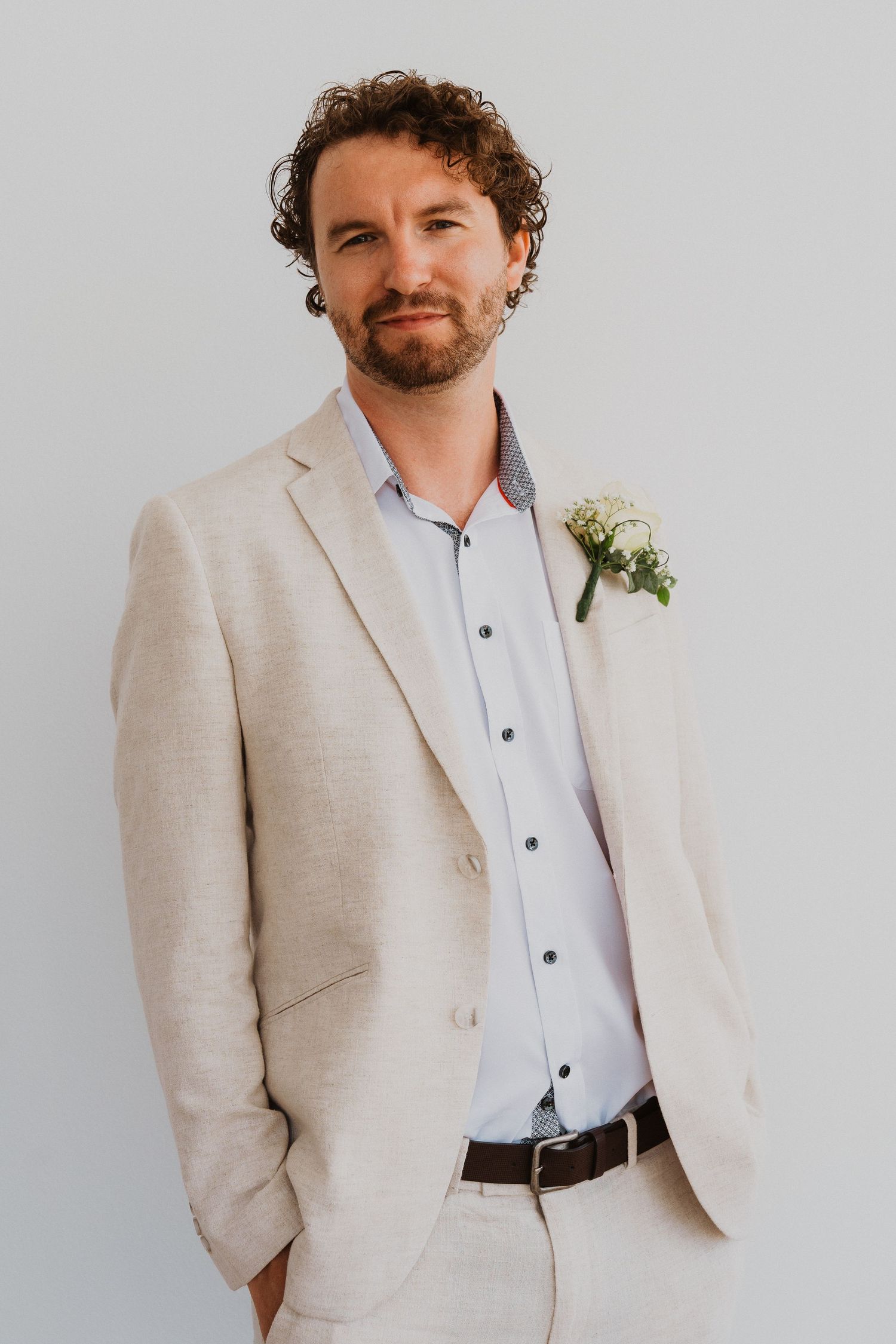 Portrait of groom in light beige suit with floral boutonniere against white background.