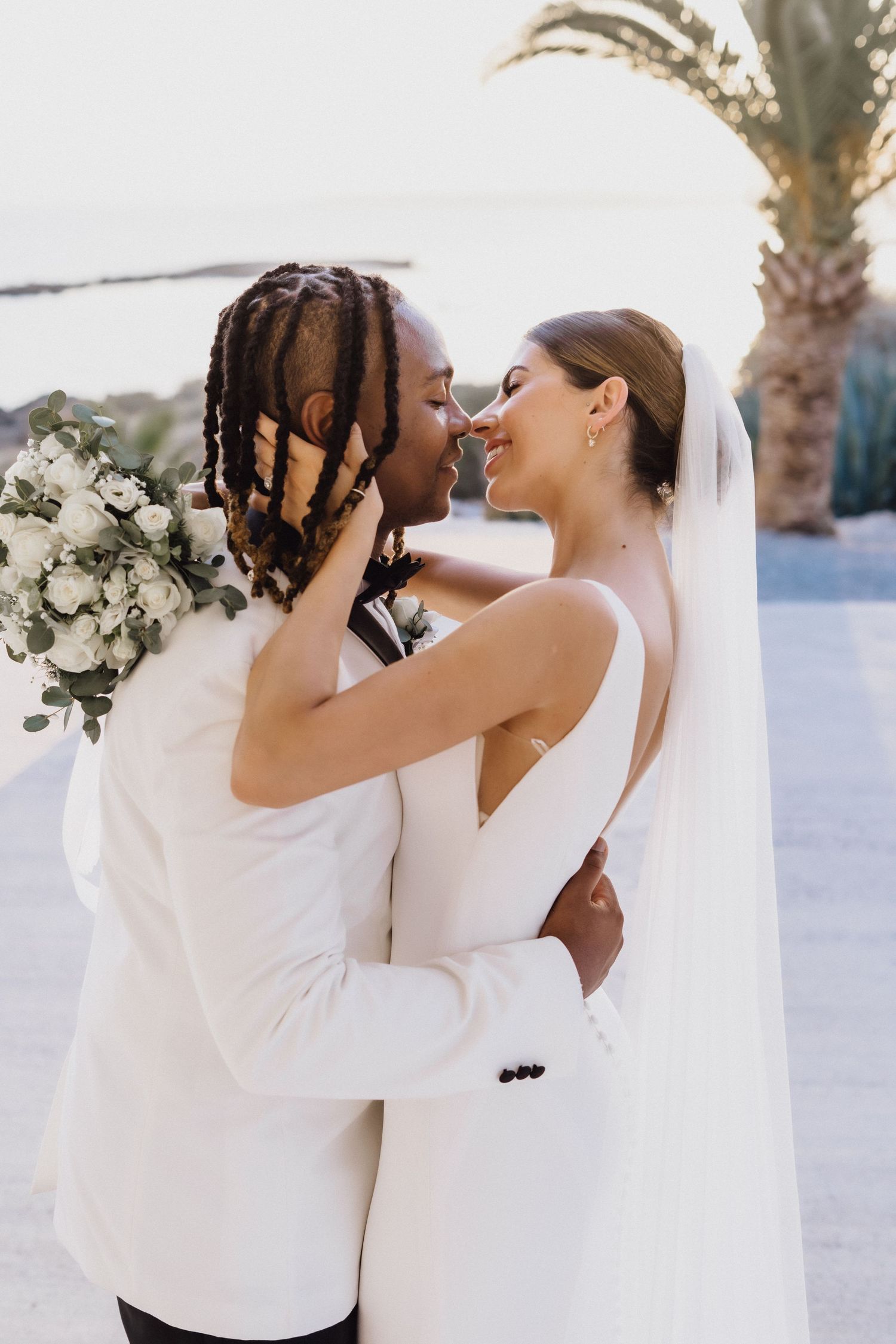 A tender wedding kiss photographed against a backdrop of palm trees and ocean views.