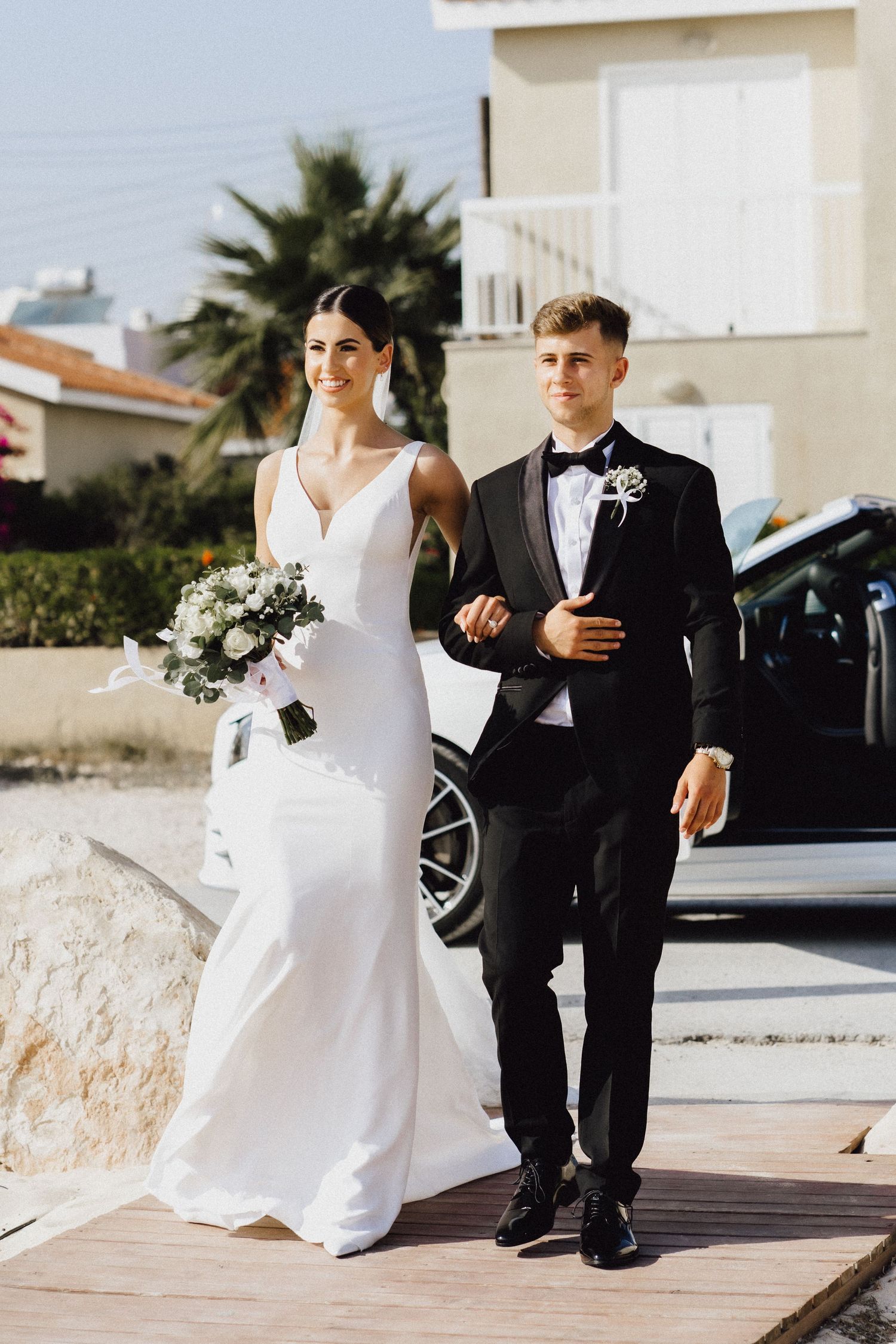 A couple walks together on a sunny day, wearing formal wedding attire near palm trees.