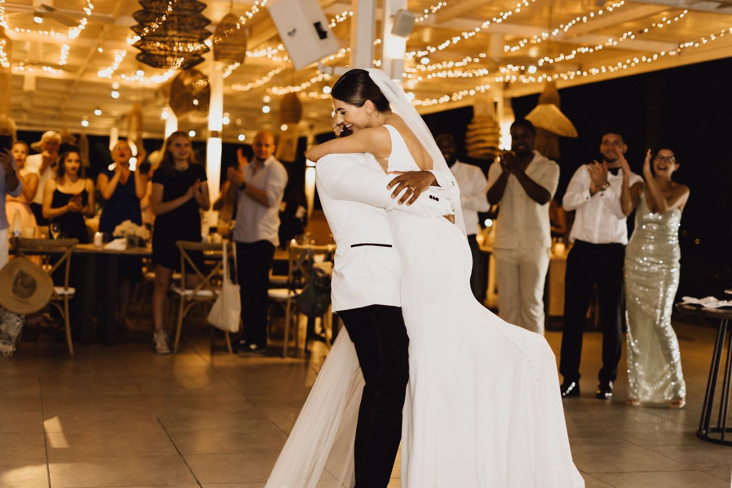 Newlyweds dance together at their wedding reception under warm string lights.