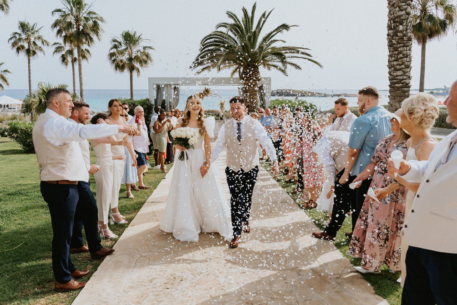 Wedding guests shower newlyweds with confetti on palm tree-lined pathway during outdoor celebration by the beach.