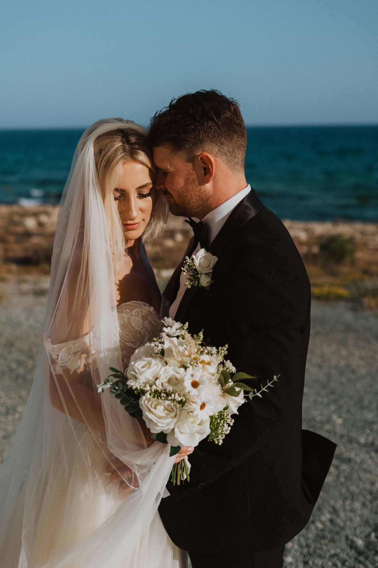 Romantic wedding couple embraces by the ocean with white floral bouquet and flowing veil at sunset.