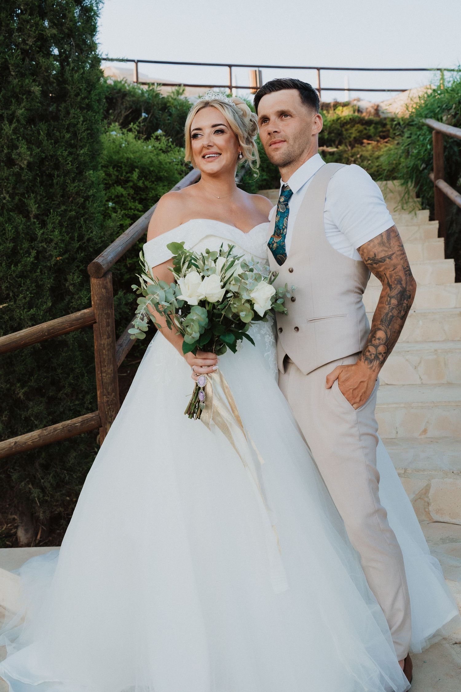 Wedding couple poses on stone steps with white floral bouquet against garden backdrop.