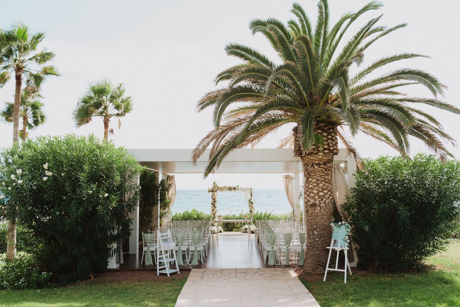 Outdoor wedding ceremony setup with white chairs and archway under tall palm trees by the ocean.