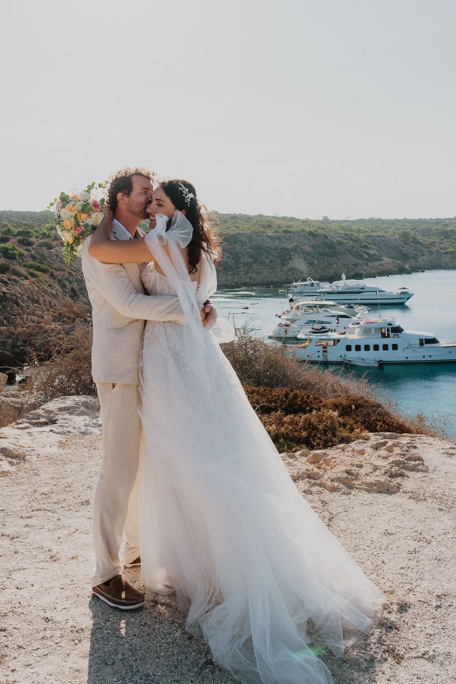 A joyful wedding couple shares an intimate moment on a cliff overlooking a harbor with yachts.
