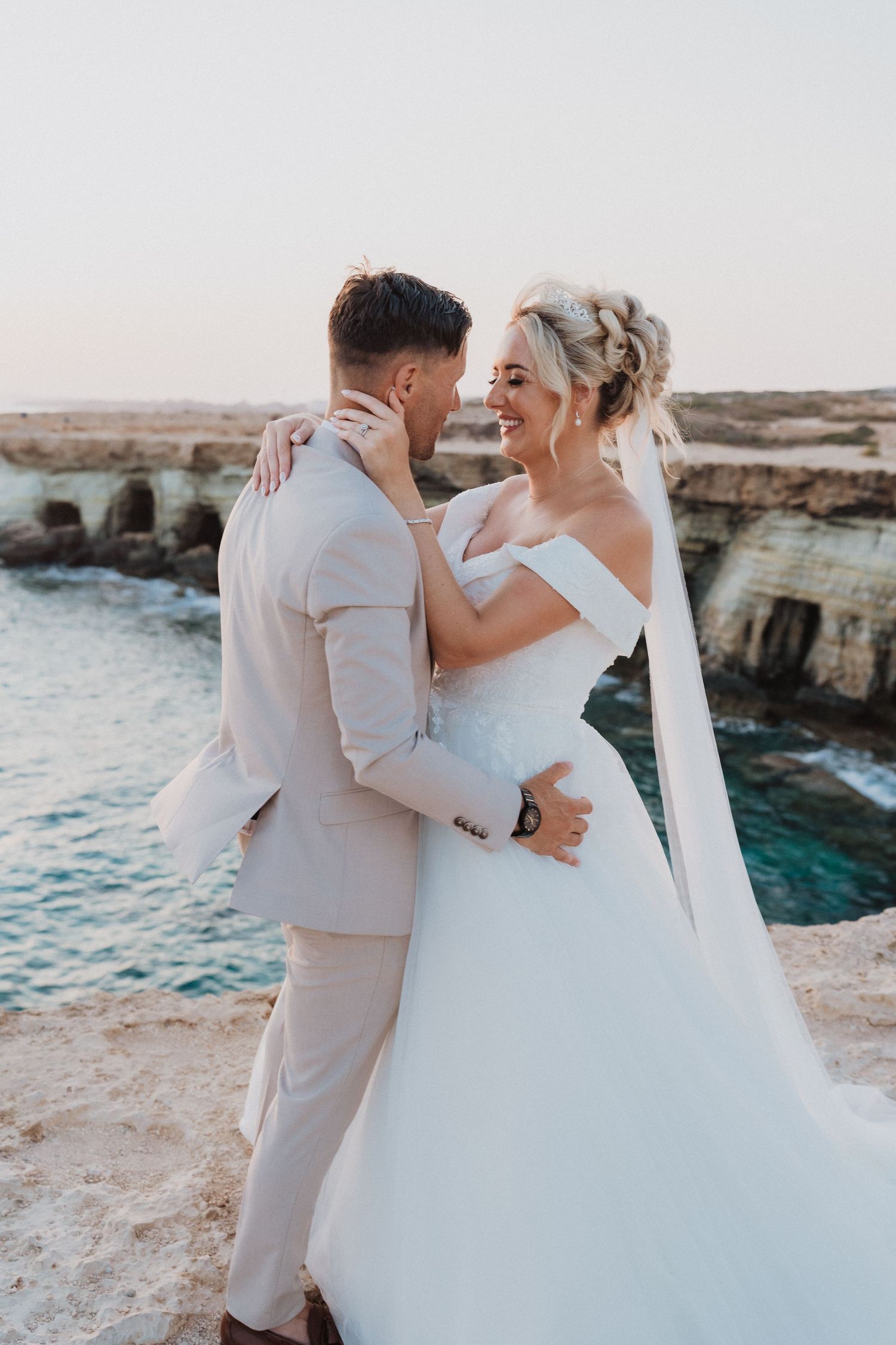 Newlywed couple sharing an intimate moment on rocky cliffs overlooking turquoise ocean waters at sunset.