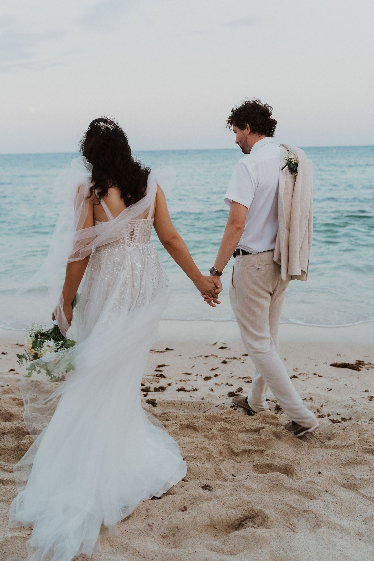 A couple walks along the shoreline holding hands during their beach wedding.