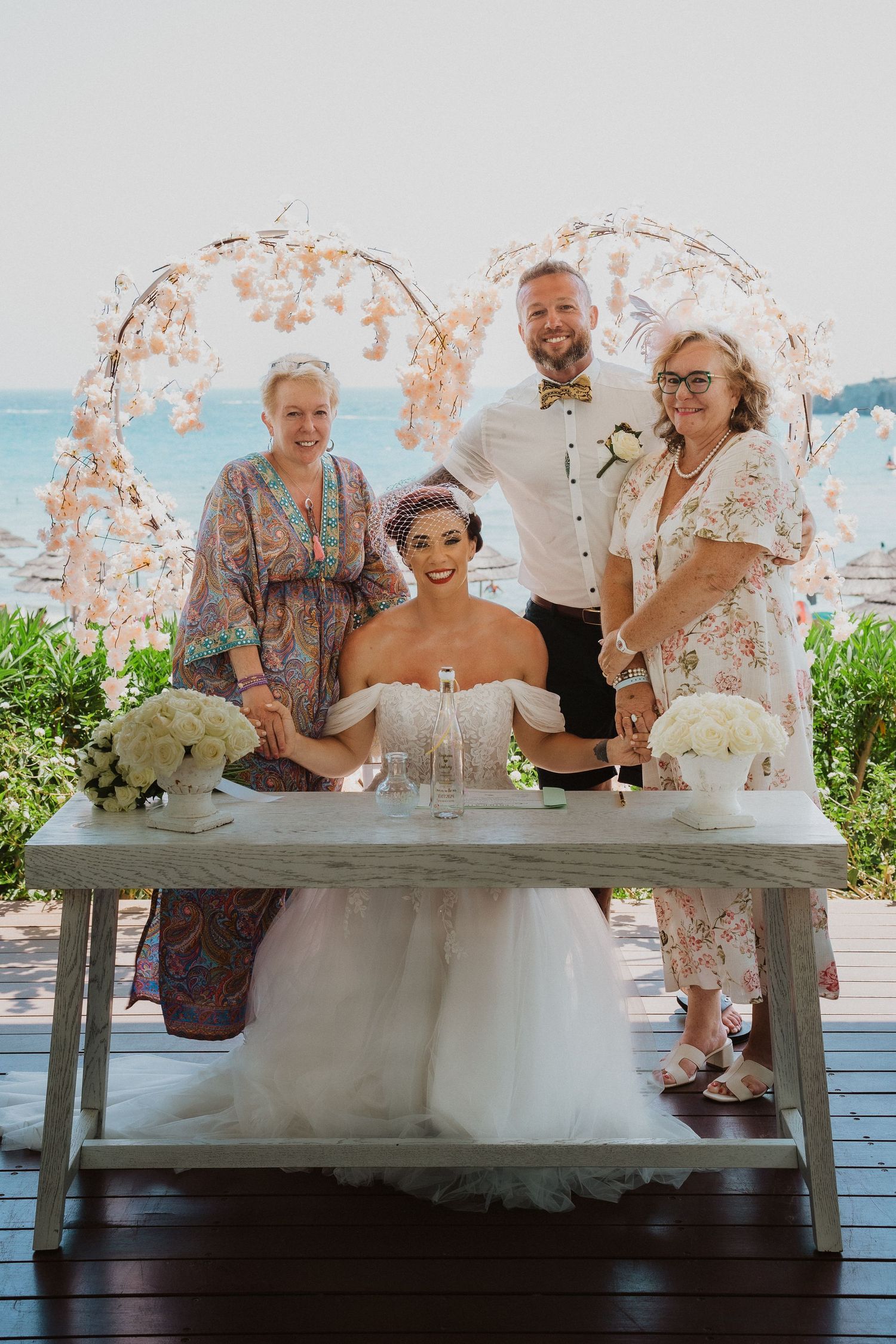 A group wedding photo at a seaside venue with floral arch and white table decorations.