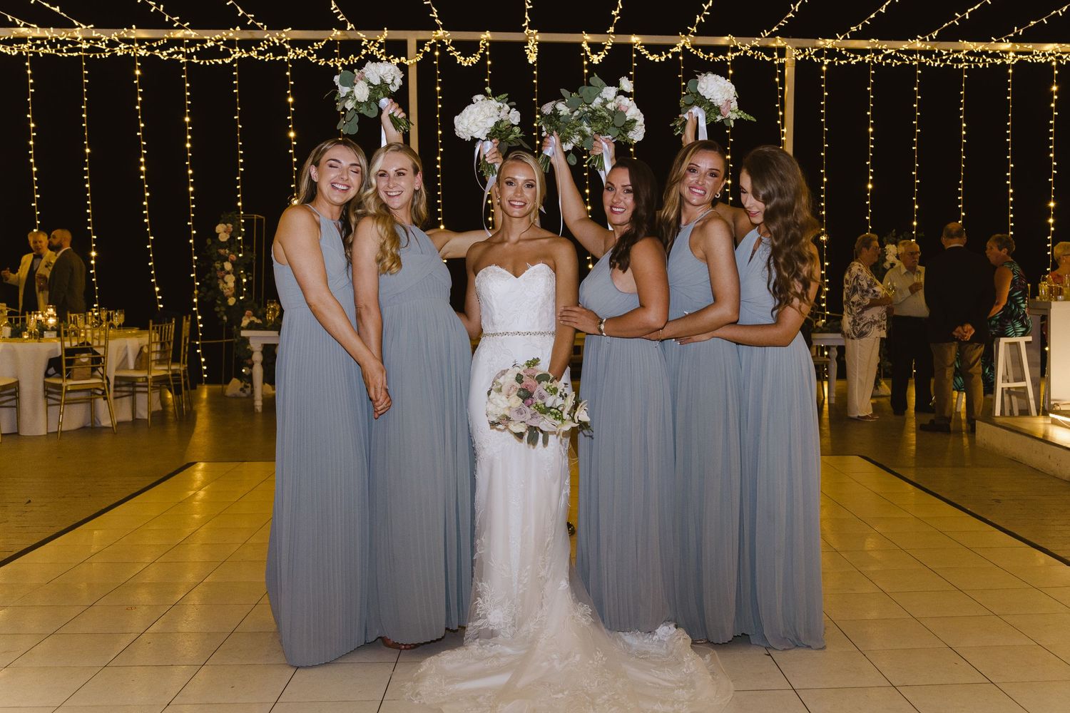 Bride in white gown poses with bridesmaids in grey dresses under string lights.