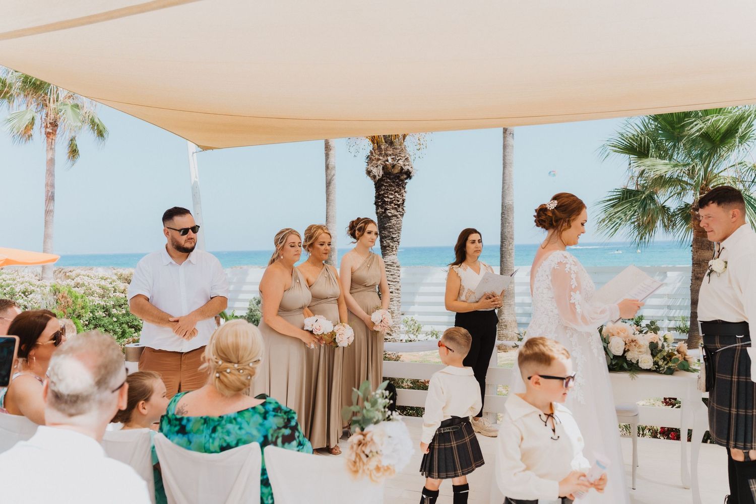 A beachside wedding ceremony features traditional Scottish attire and decor under a white tent.