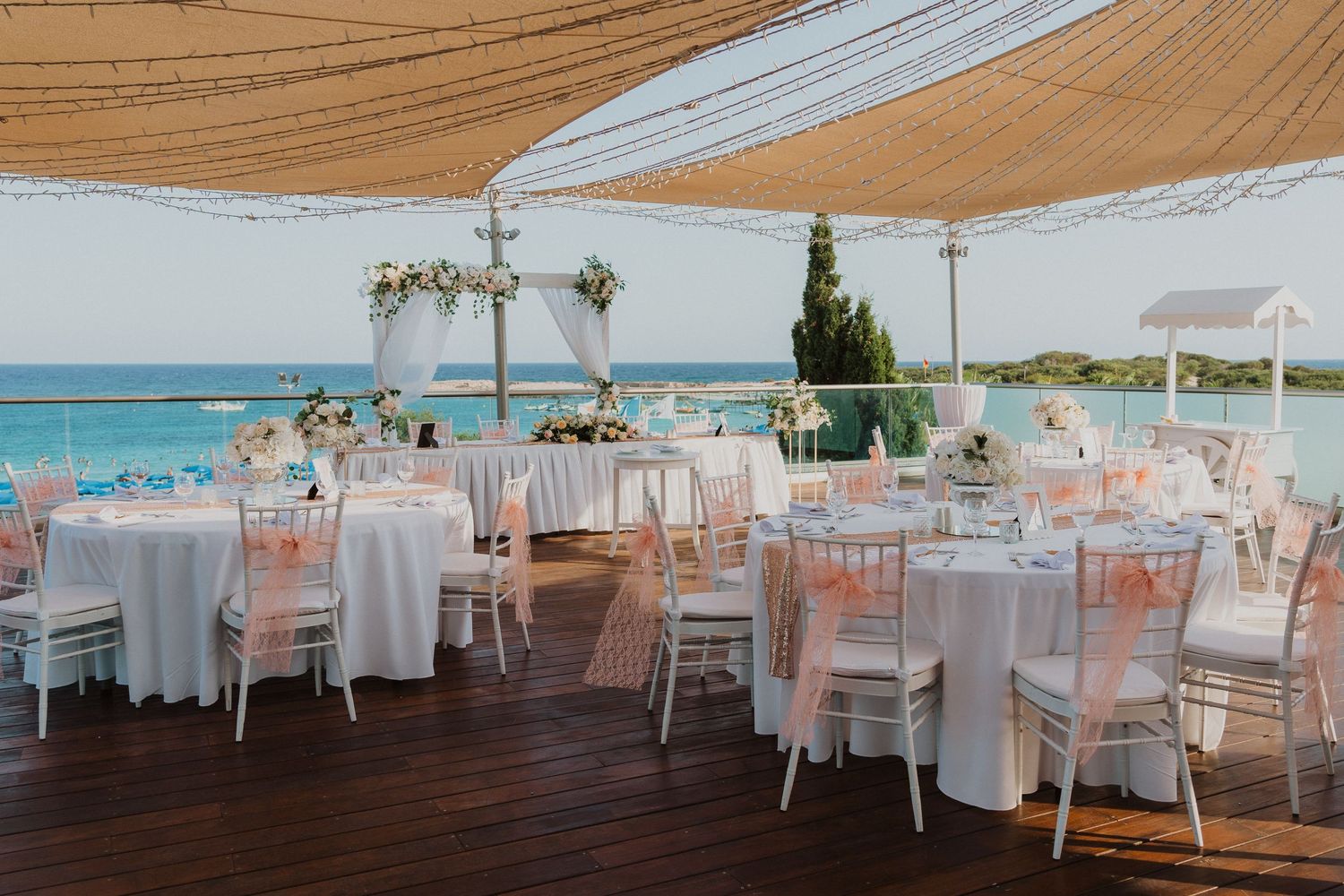 Elegant beachside wedding reception setup with white tables and pink accents under a draped canopy overlooking turquoise waters.