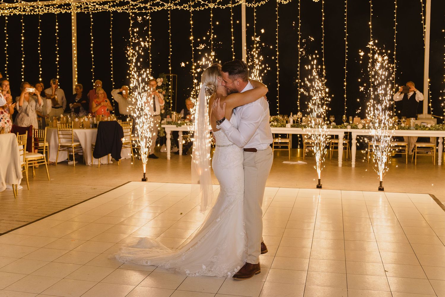 Wedding couple dances beneath golden string lights as sparklers create magical atmosphere on dance floor.