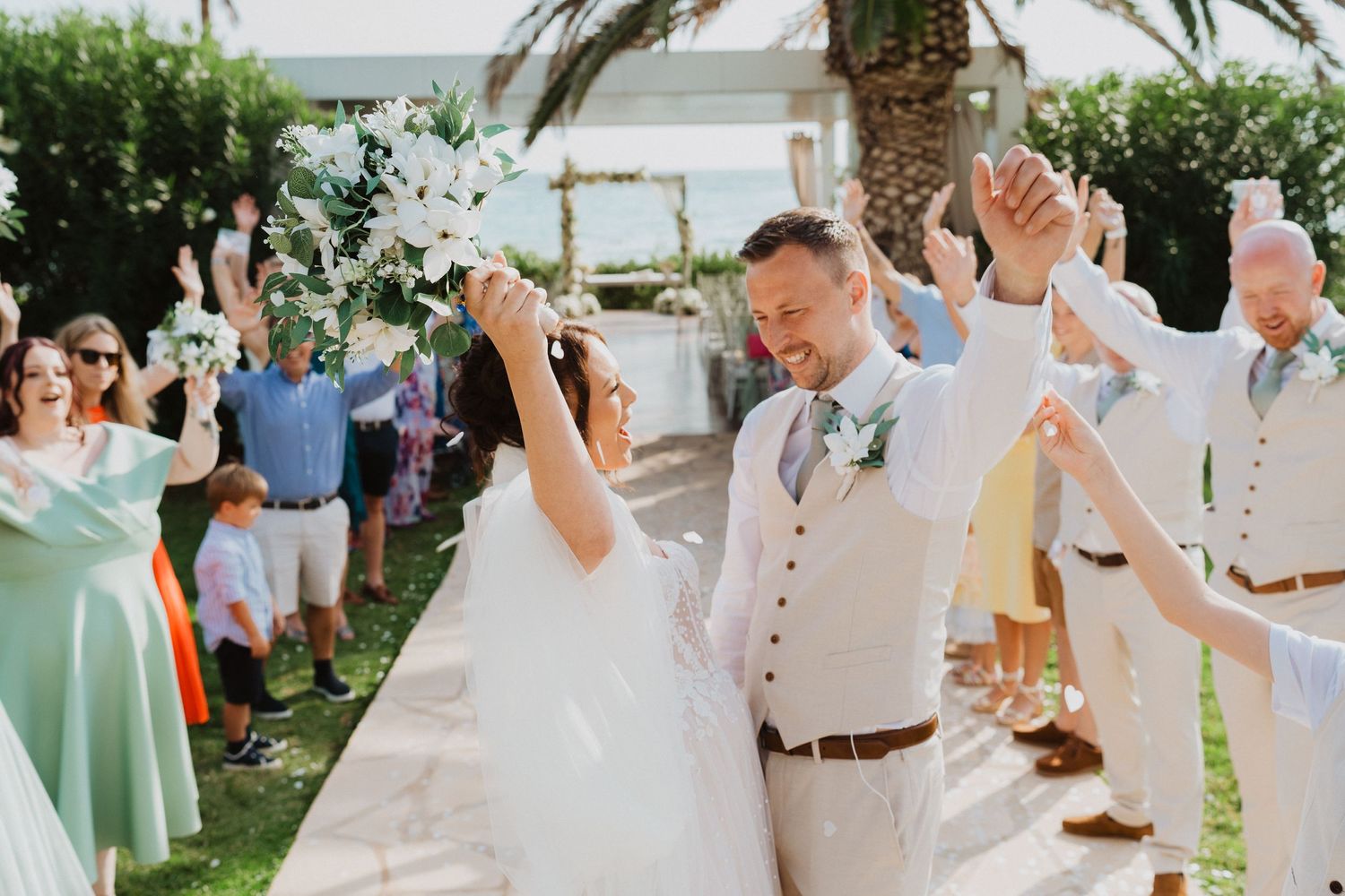 Joyful celebration at an outdoor garden wedding with guests in mint green dresses and light colored suits under palm trees.