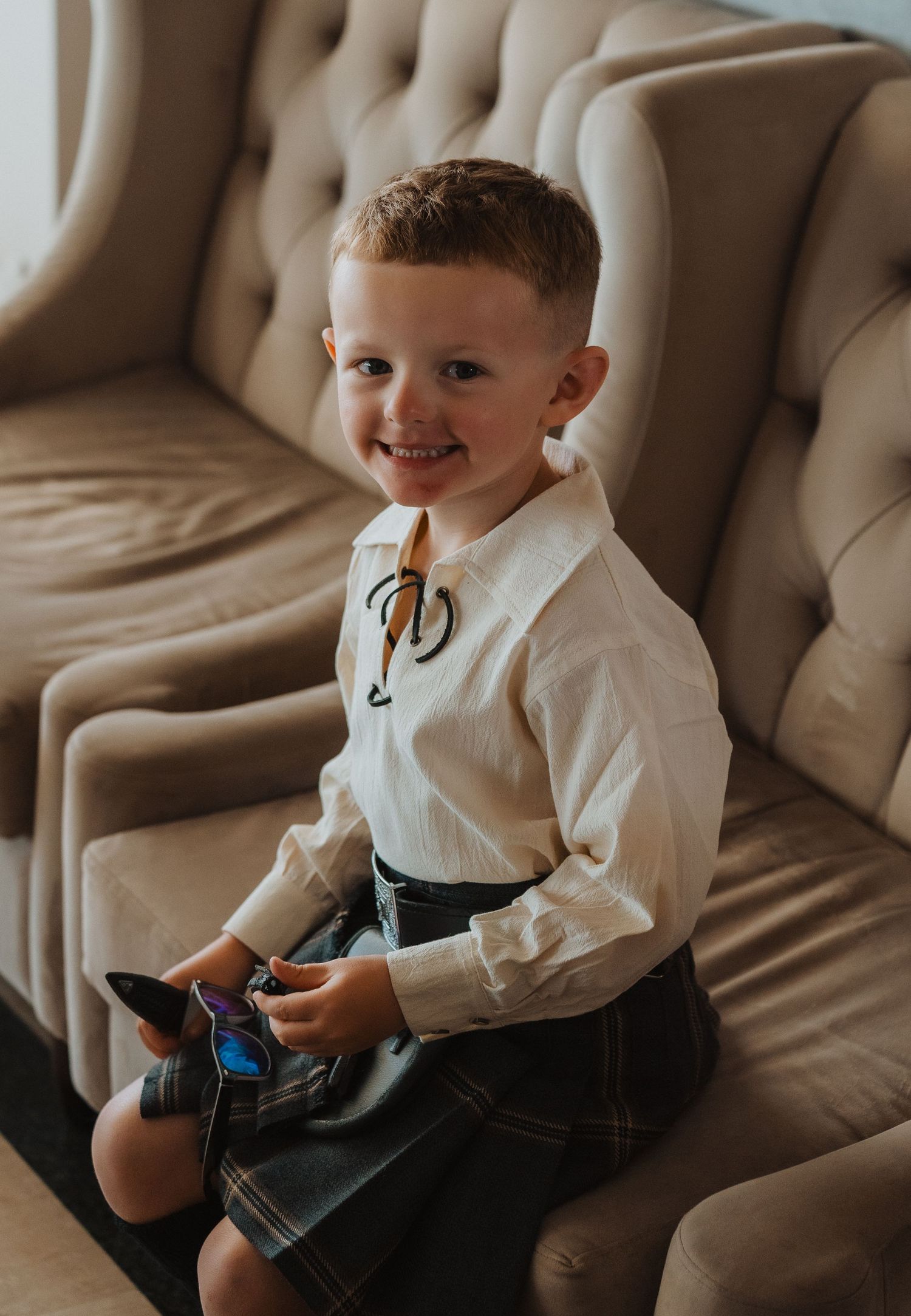 A young boy wearing traditional Scottish attire with a white shirt and kilt smiles while sitting on a beige couch.
