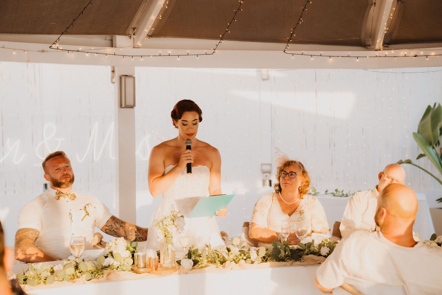 Wedding reception speech moment with decorated head table and white florals.