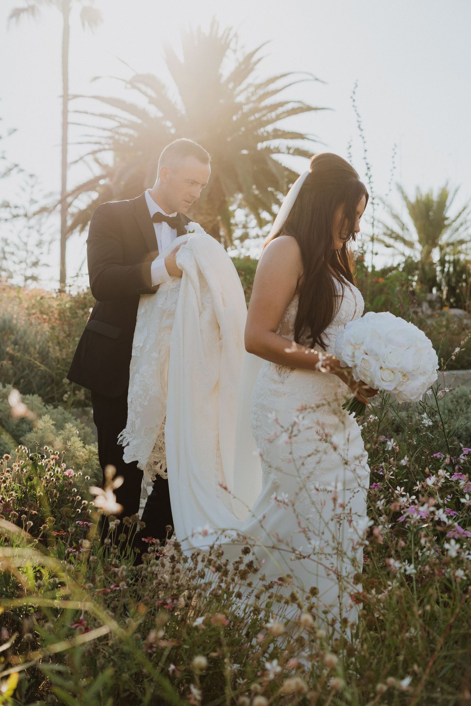 A couple in formal wedding attire stands among wildflowers and palm trees during golden hour.
